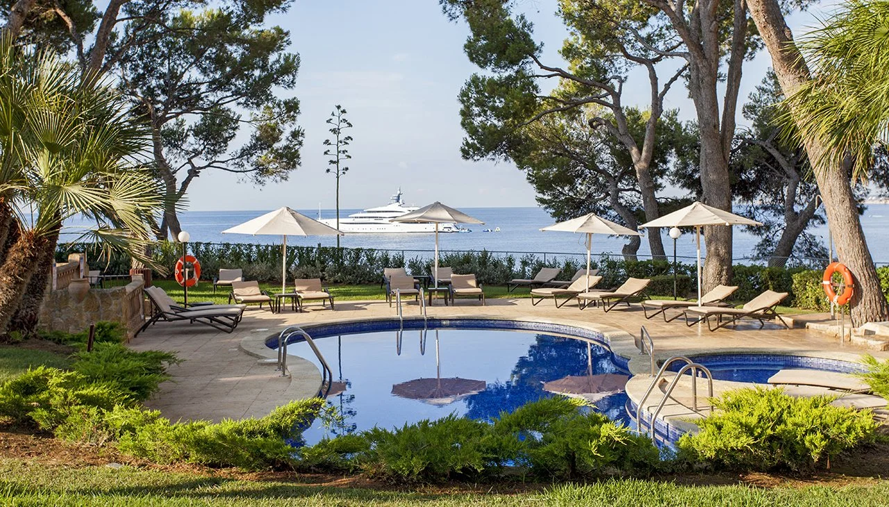 Piscina en un área al aire libre con sillas y sombrillas, vista al mar con yate y árboles alrededor.