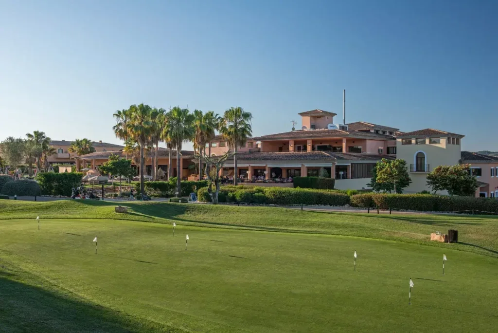 Vista de un campo de golf con un edificio grande y palmeras en el fondo, en un día soleado.