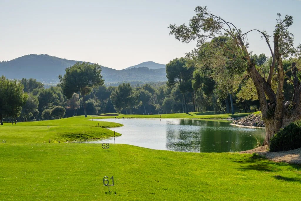 Vista de un campo de golf con césped bien cuidado, un lago y árboles, con montañas al fondo bajo un cielo despejado.