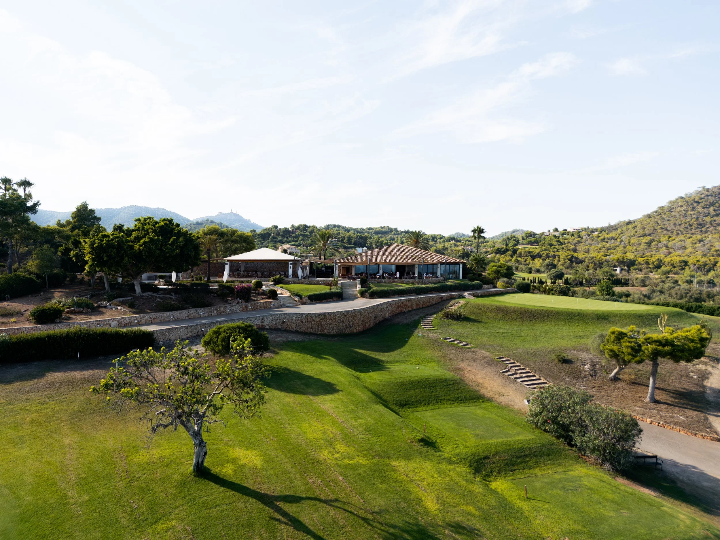Vista panorámica de un campo de golf con césped cuidado, árboles verdes dispersos, una estructura en el fondo y colinas en el horizonte bajo un cielo soleado.