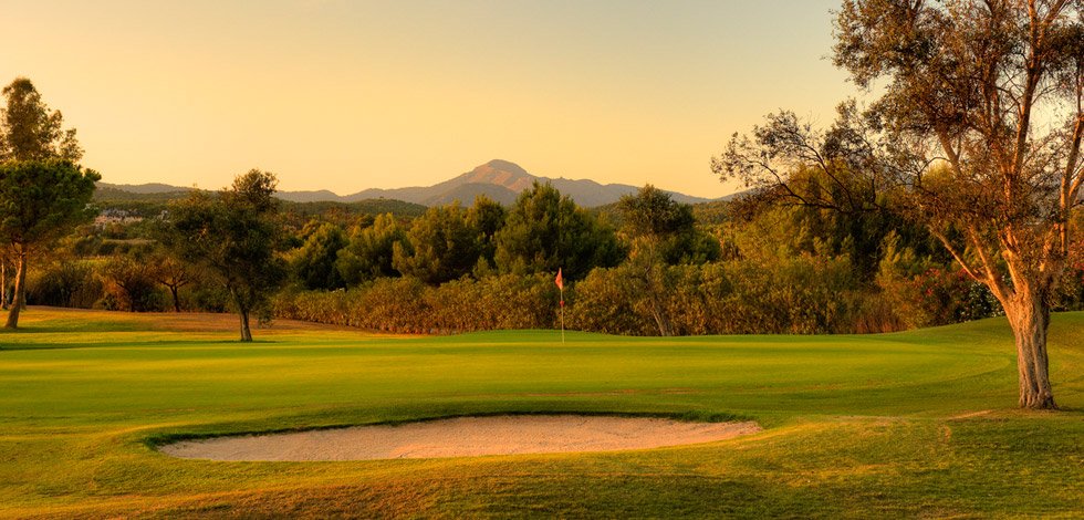 Campo de golf con greens, un hoyo y árboles, al atardecer con montañas de fondo.