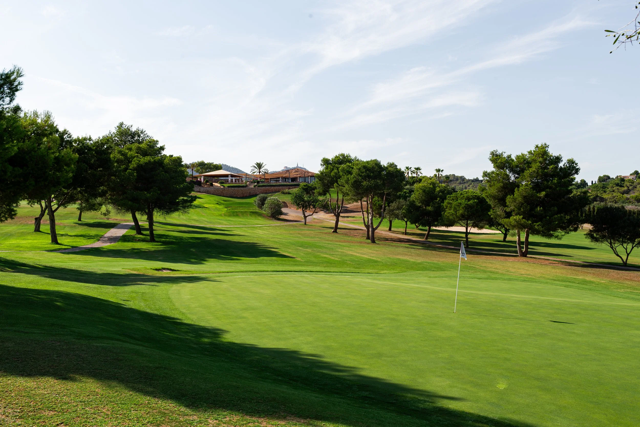 Un campo de golf bien cuidado con árboles dispersos y un área de césped con bandera, en un día soleado con cielo despejado.