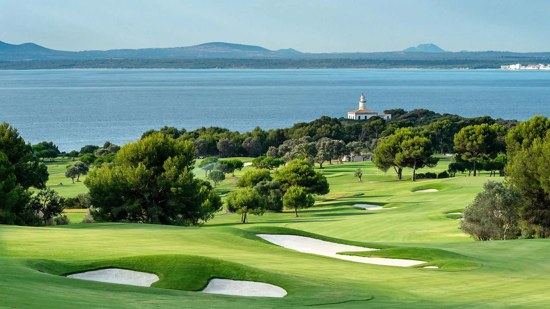 Campo de golf bien cuidado con búnkeres, árboles dispersos y vista de un lago y colinas en el fondo, con un faro en una colina cerca del agua.