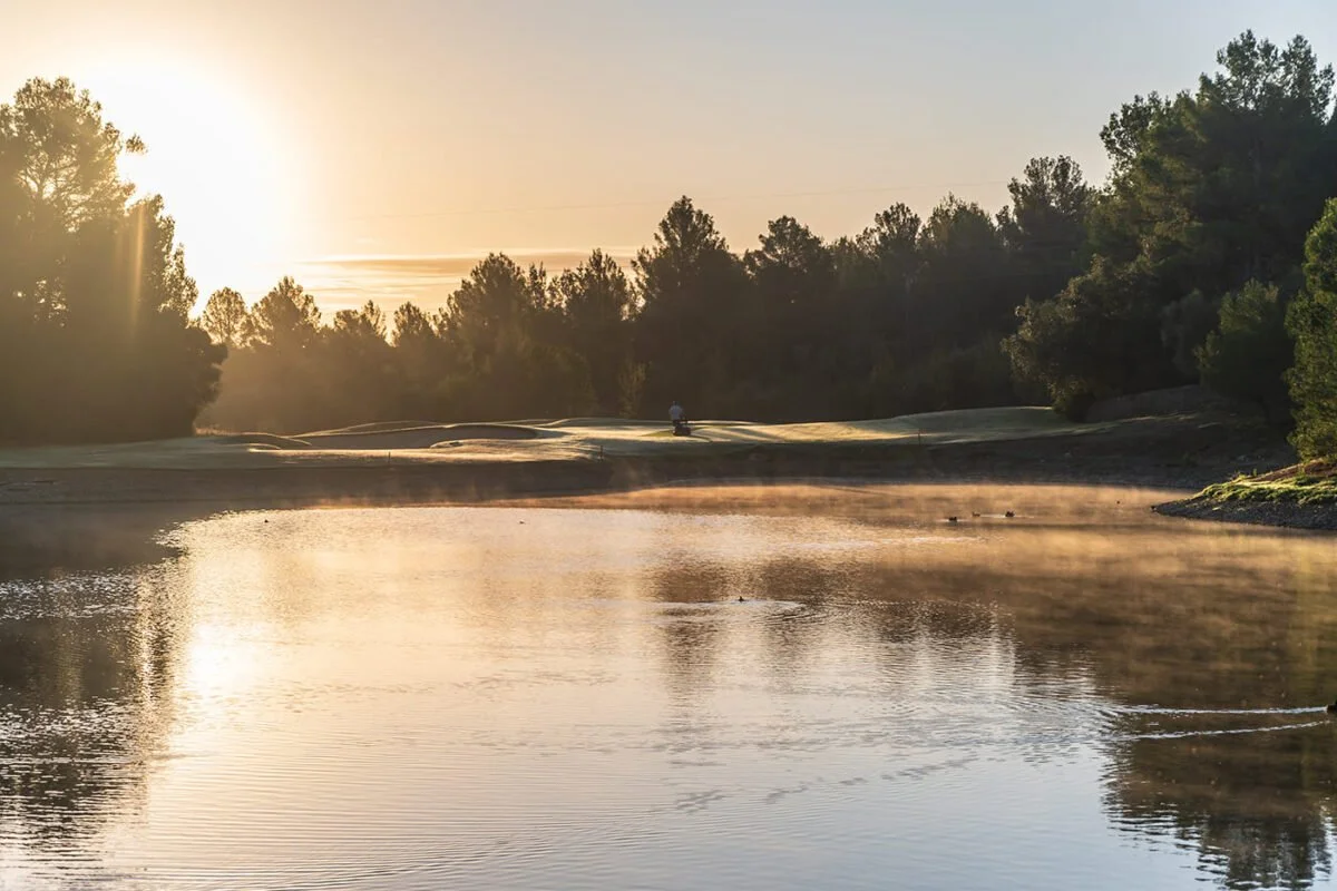 Vista de un río con vapor saliendo del agua, rodeado de árboles y una cancha de golf al atardecer.