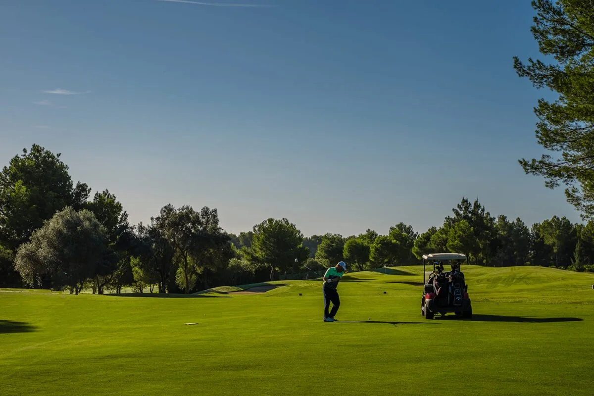 Persona jugando golf en un campo con un carrito de golf y árboles en el fondo.