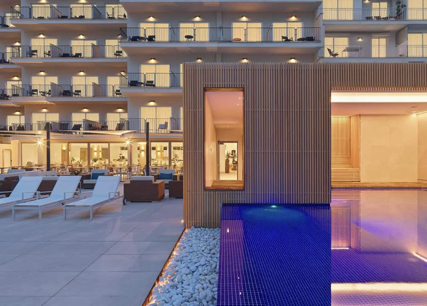 Vista de un hotel de lujo con alberca y áreas de descanso, en la noche, con iluminación cálida y moderna arquitectura.
