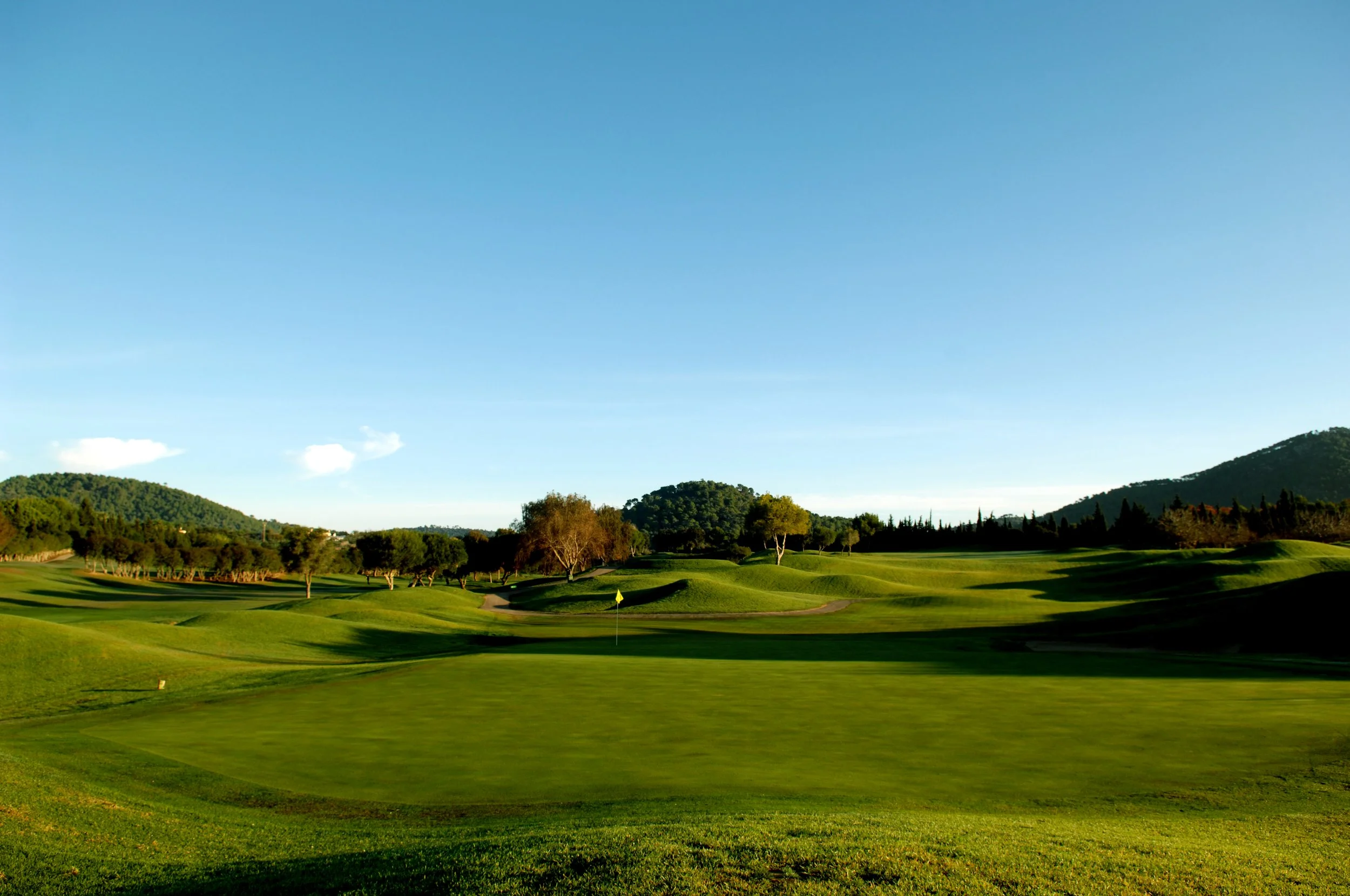 Campo de golf con césped verde, árboles dispersos y colinas al fondo bajo un cielo despejado.