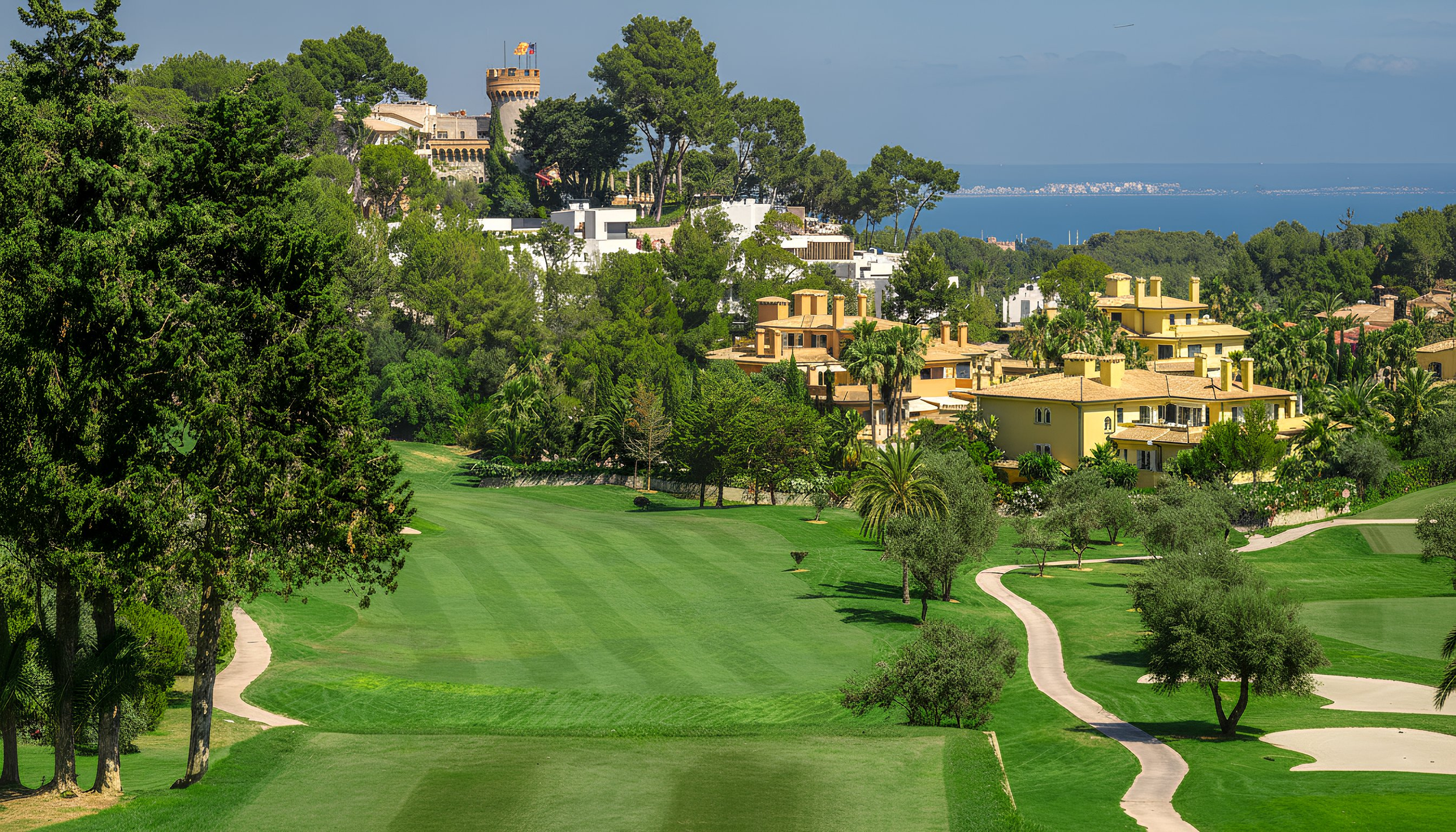 Un campo de golf con césped bien cuidado, árboles y caminos serpenteantes, con casas residenciales en las colinas y vista al mar al fondo, bajo un cielo despejado.