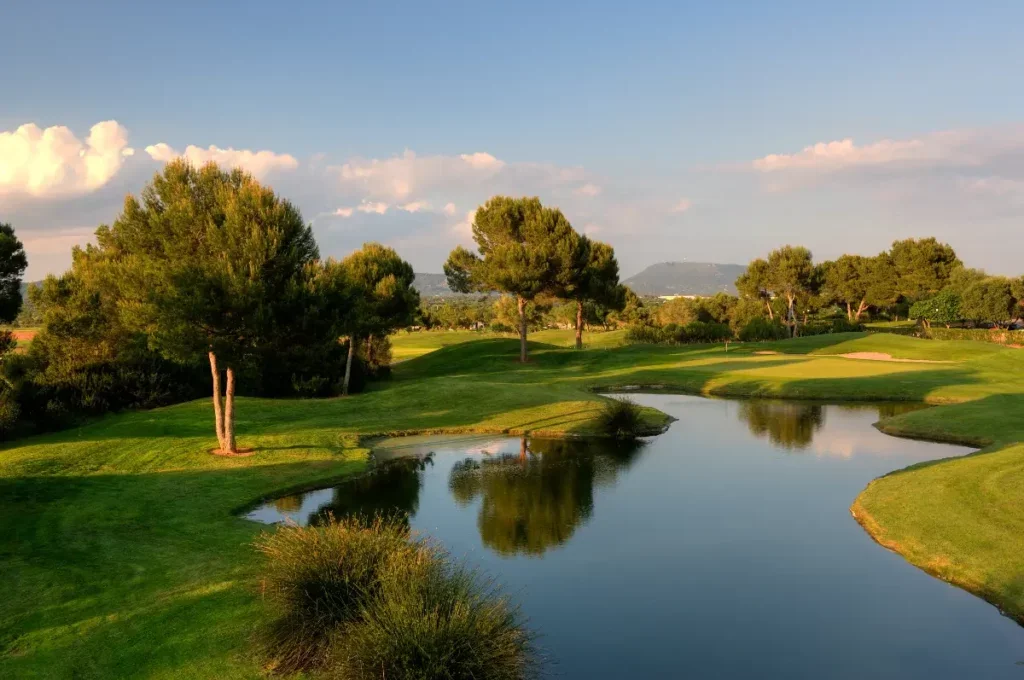 Campo de golf con árboles y un pequeño lago, cielo despejado con nubes.
