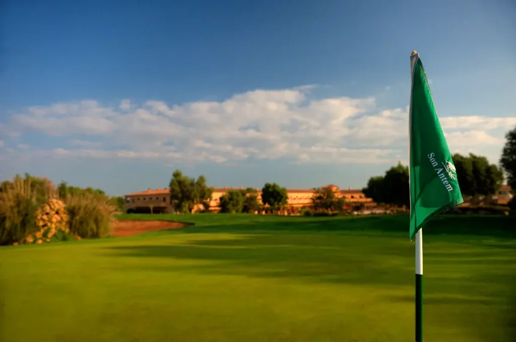 Vista de un campo de golf con una bandera verde en primer plano, árboles y edificaciones al fondo bajo un cielo parcialmente nublado.