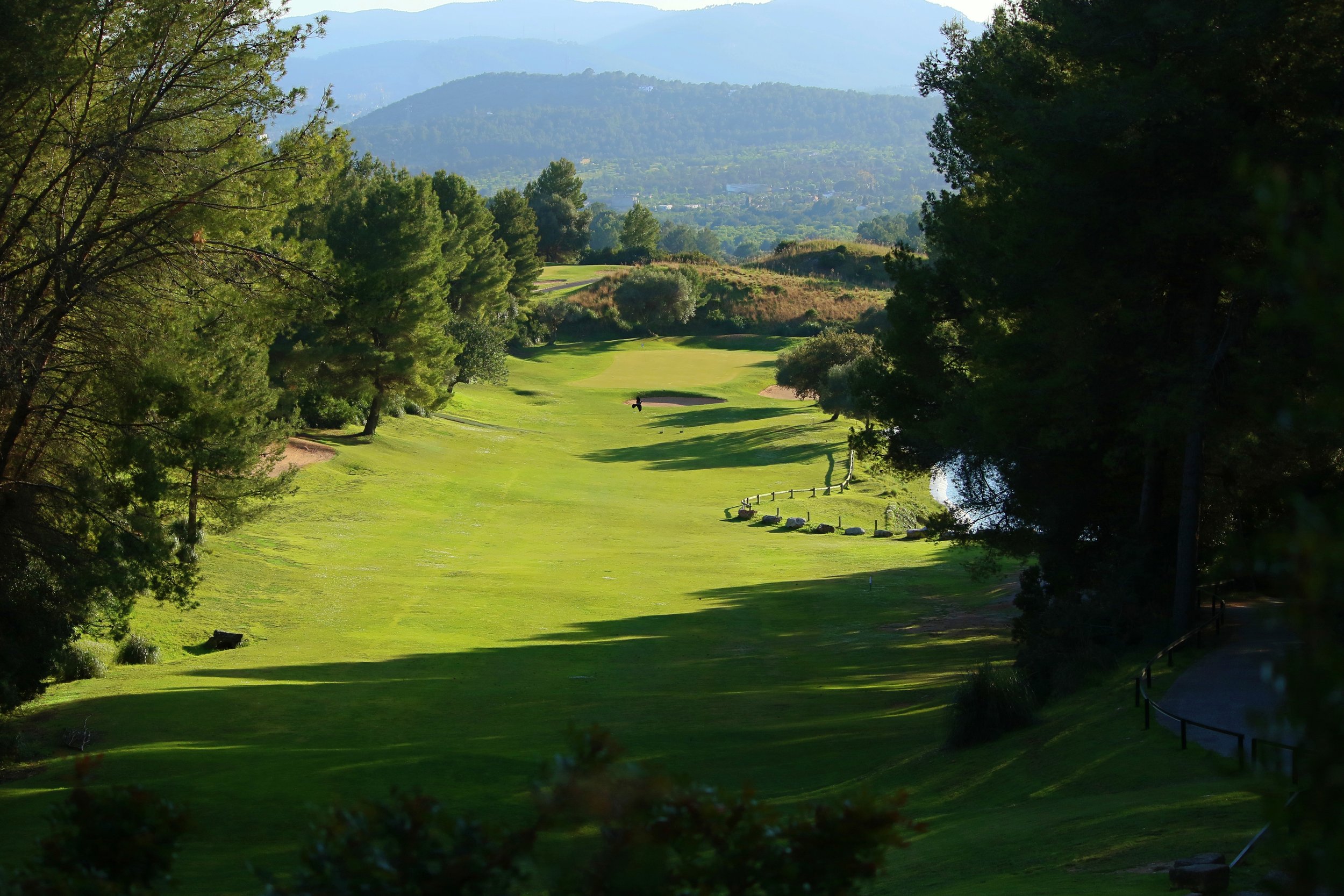 Un campo de golf rodeado de árboles y colinas en el fondo, con un riachuelo a la derecha y sombras en el césped.