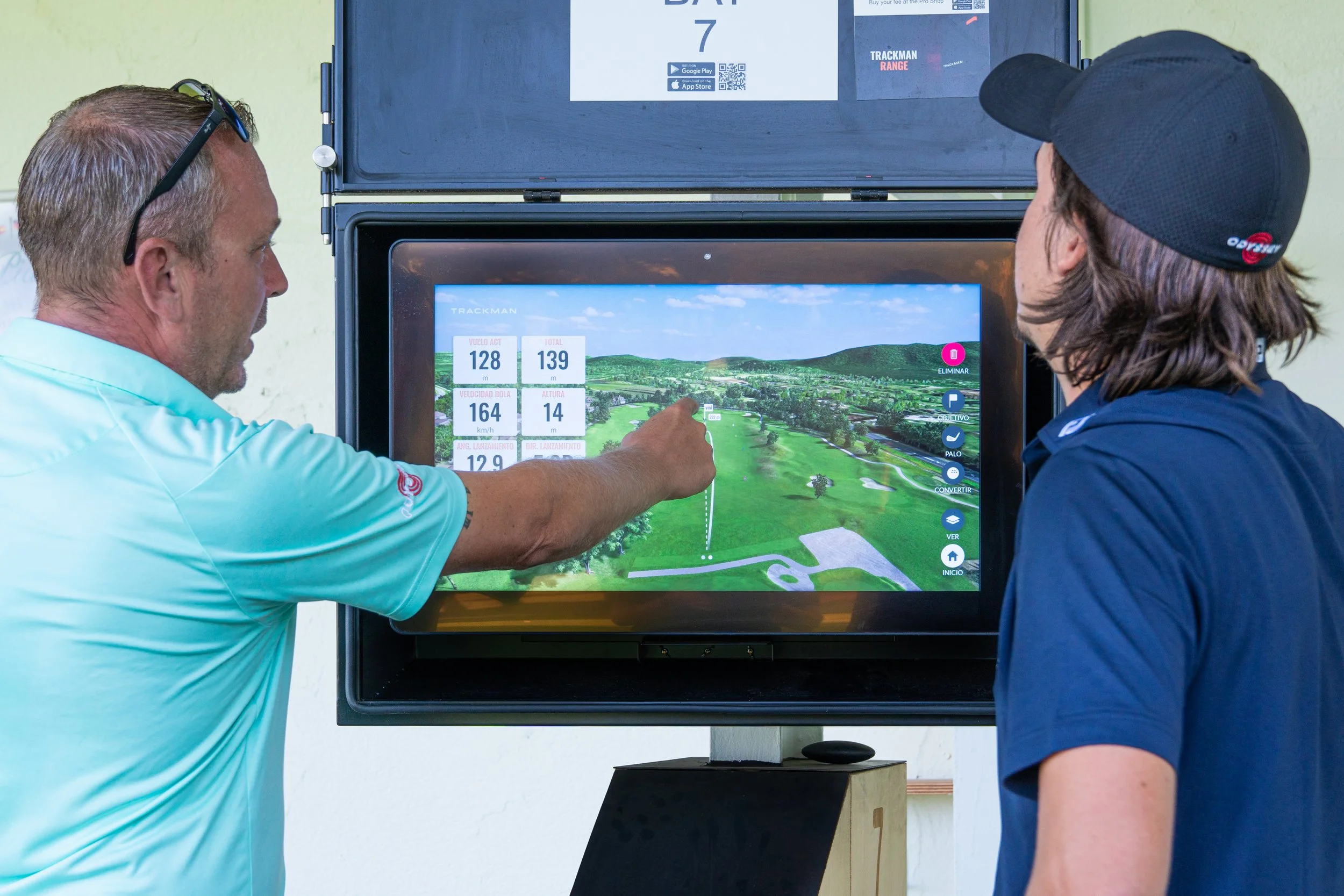 Dos hombres observando una pantalla de simulación de golf, uno muestra información de distancia y otros datos relacionados con el juego.
