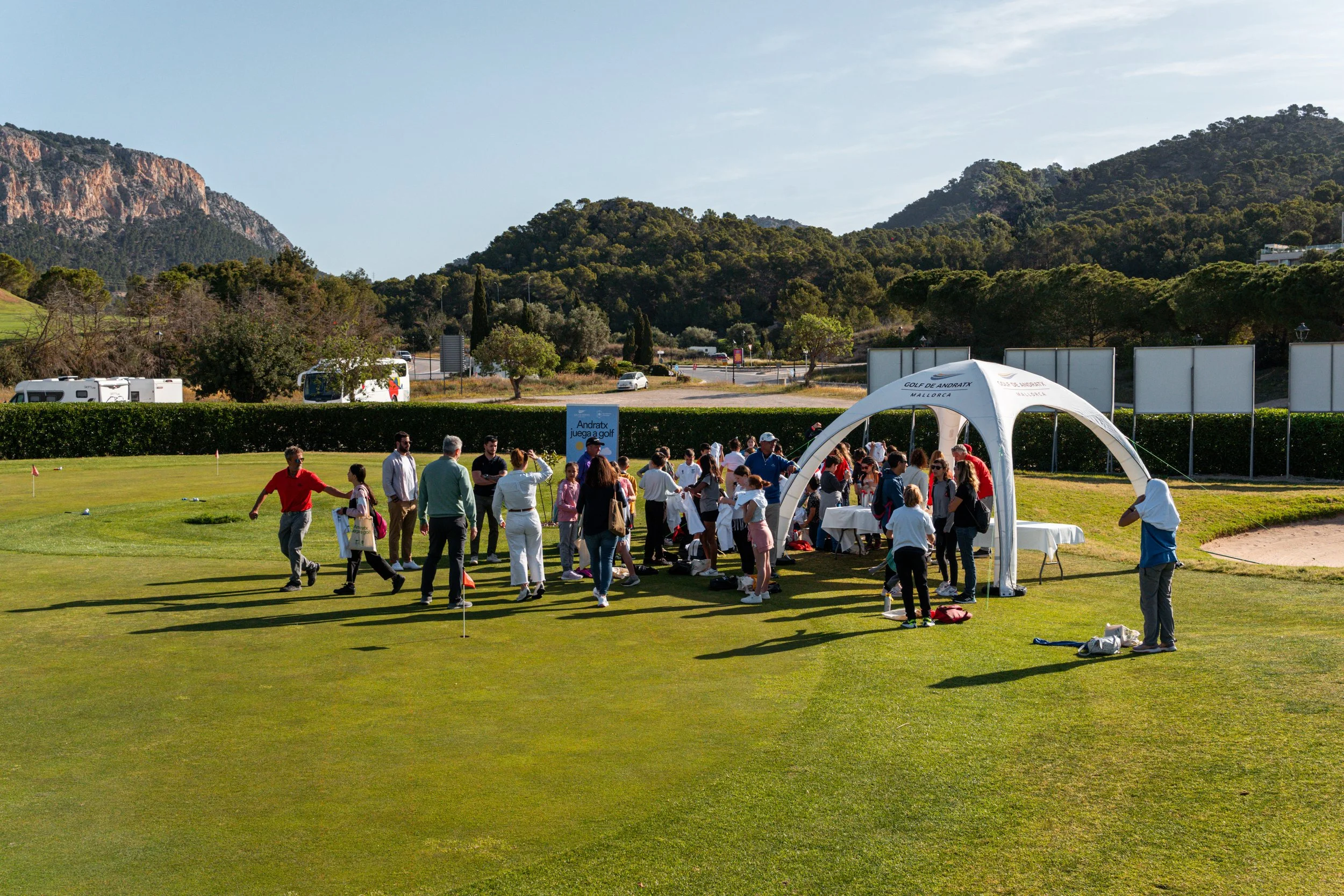 Grupo de personas en un campo de golf al aire libre, con una carpa blanca y herramientas de golf, rodeadas de montañas y árboles.