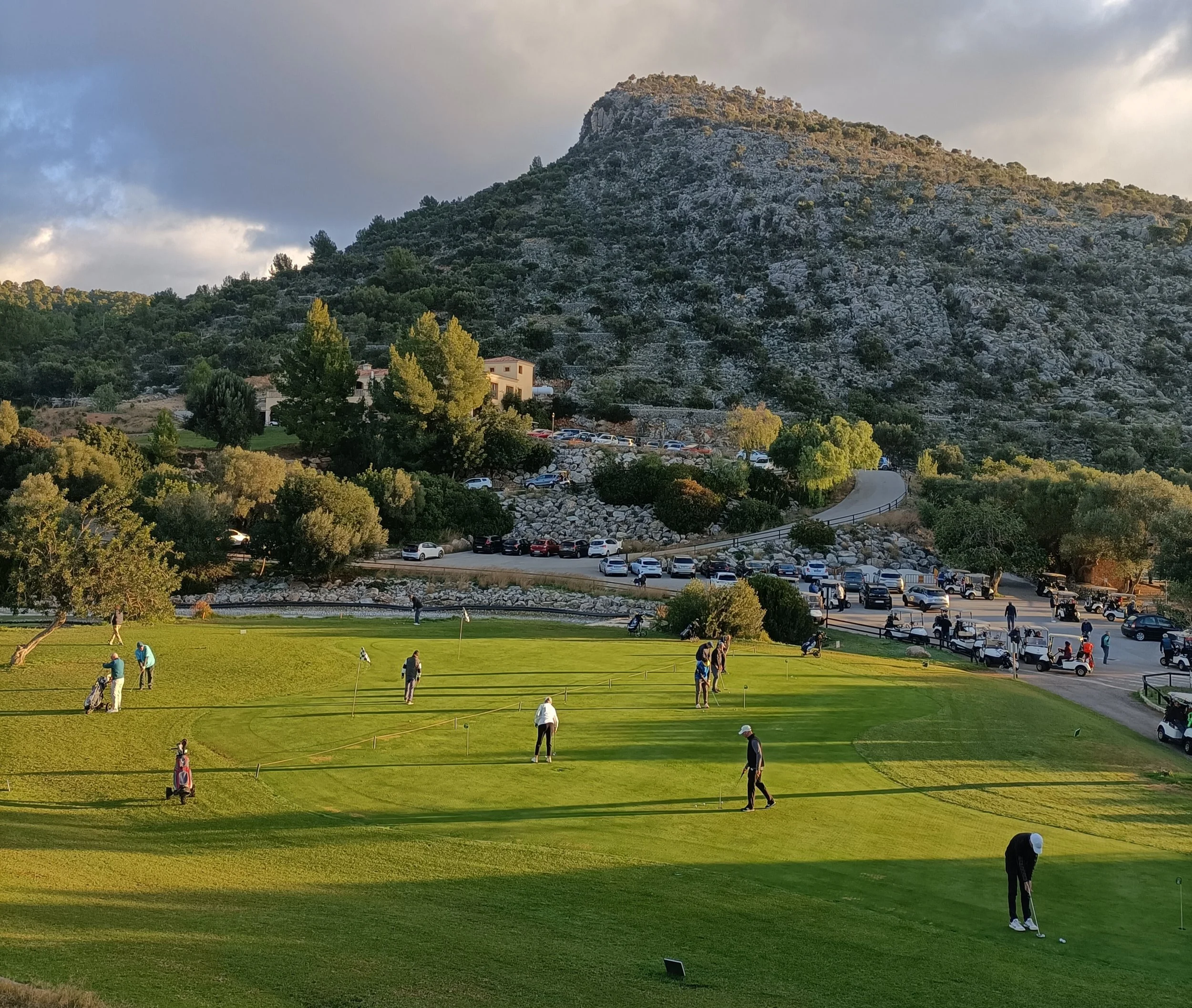 Personas jugando golf en un campo con colinas y árboles, con parqueo lleno en el fondo, montañas y cielo nublado.