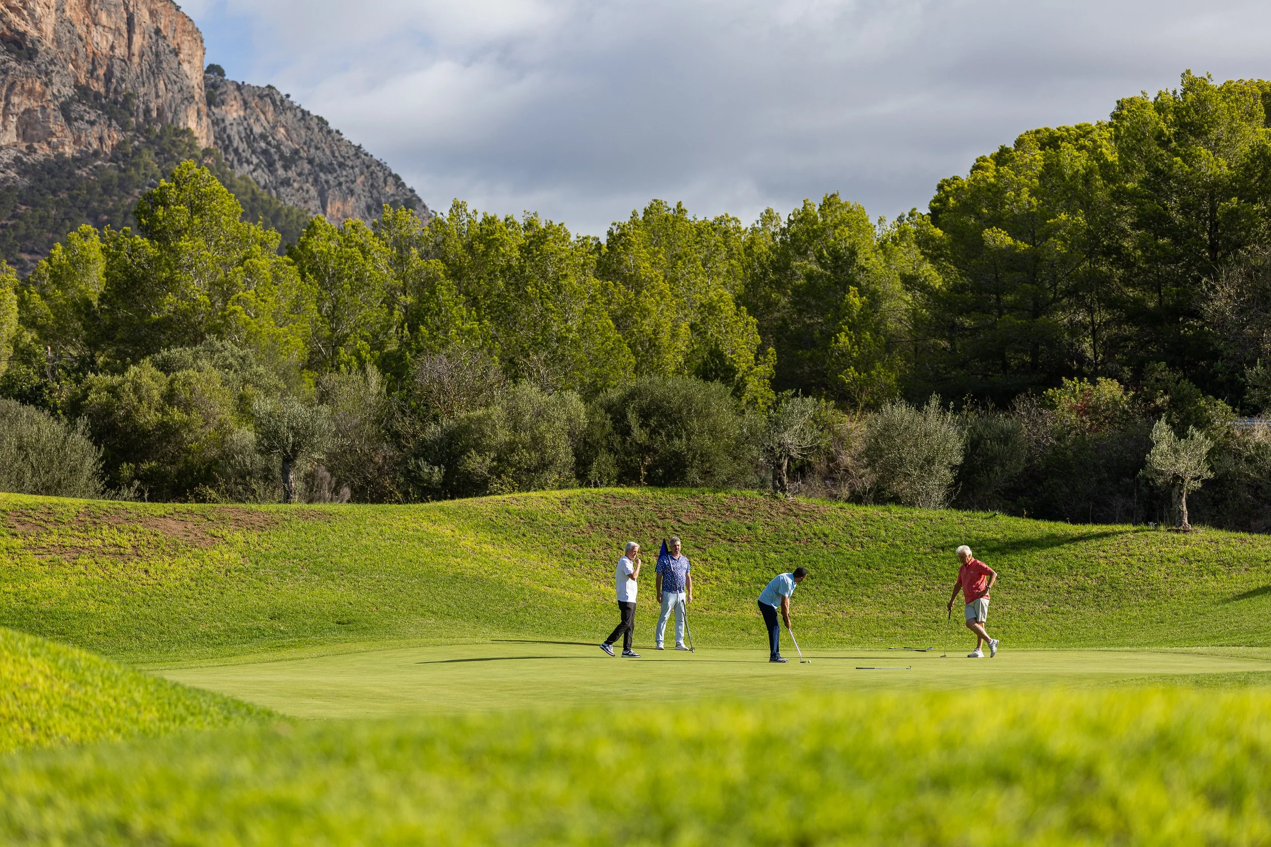 Cinco personas jugando golf en un campo verde rodeado de árboles y montañas en un día nublado.