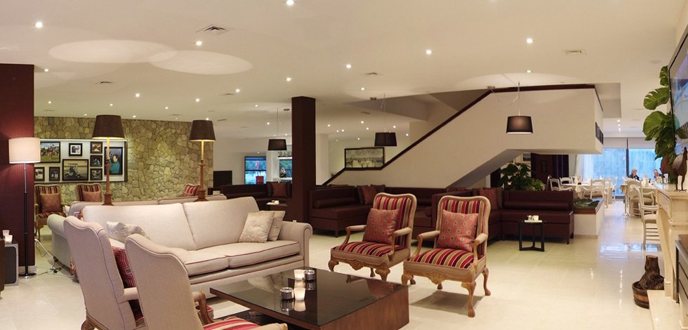 Interior de un restaurante o sala de hotel elegantemente decorada con sillones y sillas, iluminación tenue, paredes con cuadros y plantas.