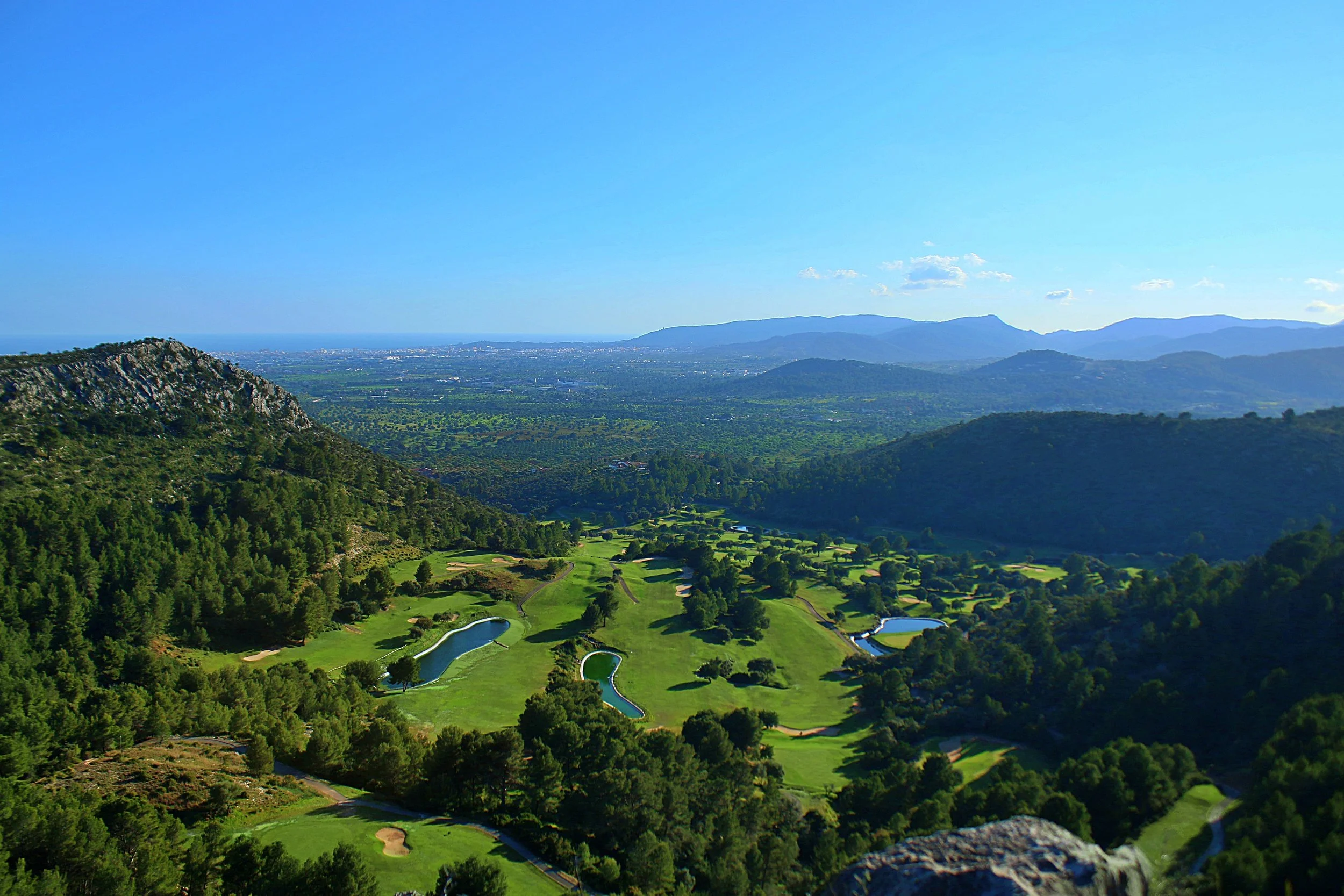 Vistas de un campo de golf rodeado de árboles y montañas en el fondo, bajo un cielo despejado.
