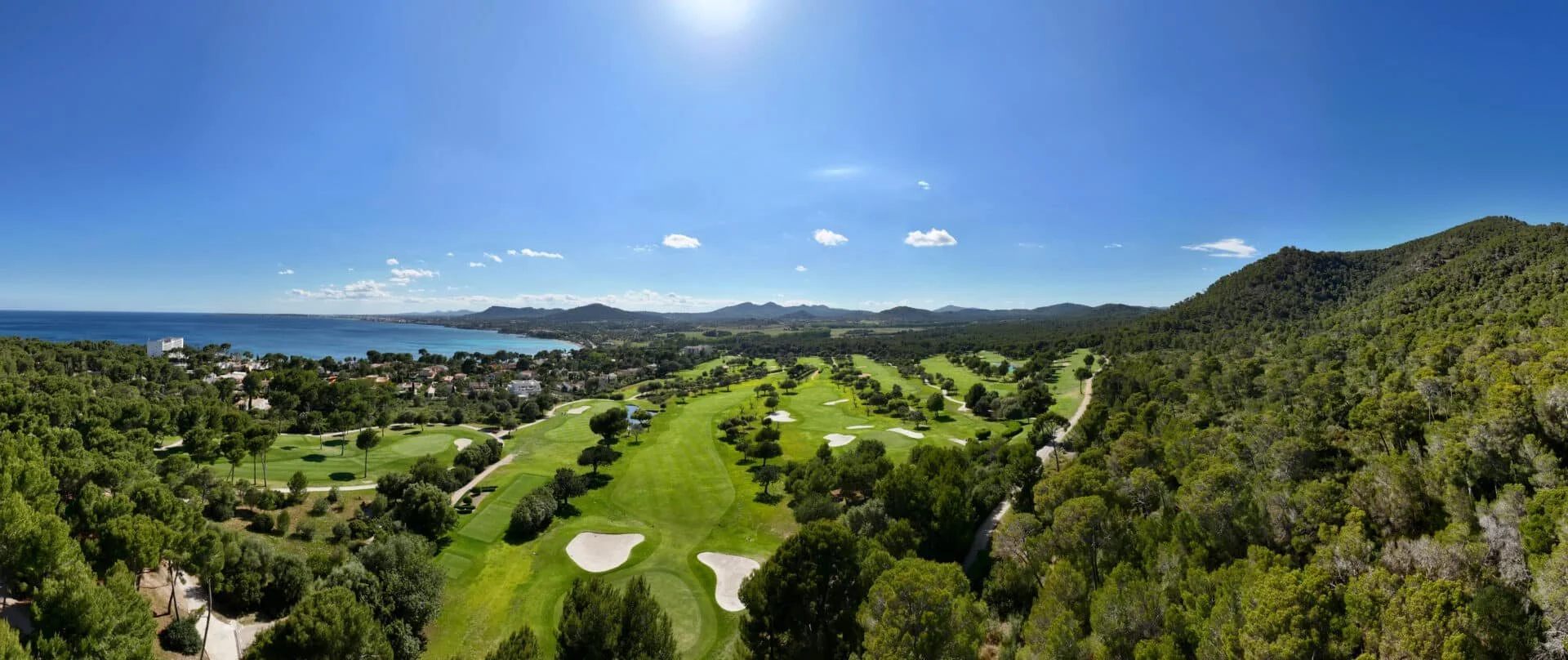 Campo de golf rodeado de árboles y cerca del mar, bajo un cielo despejado y soleado con algunas nubes pequeñas.