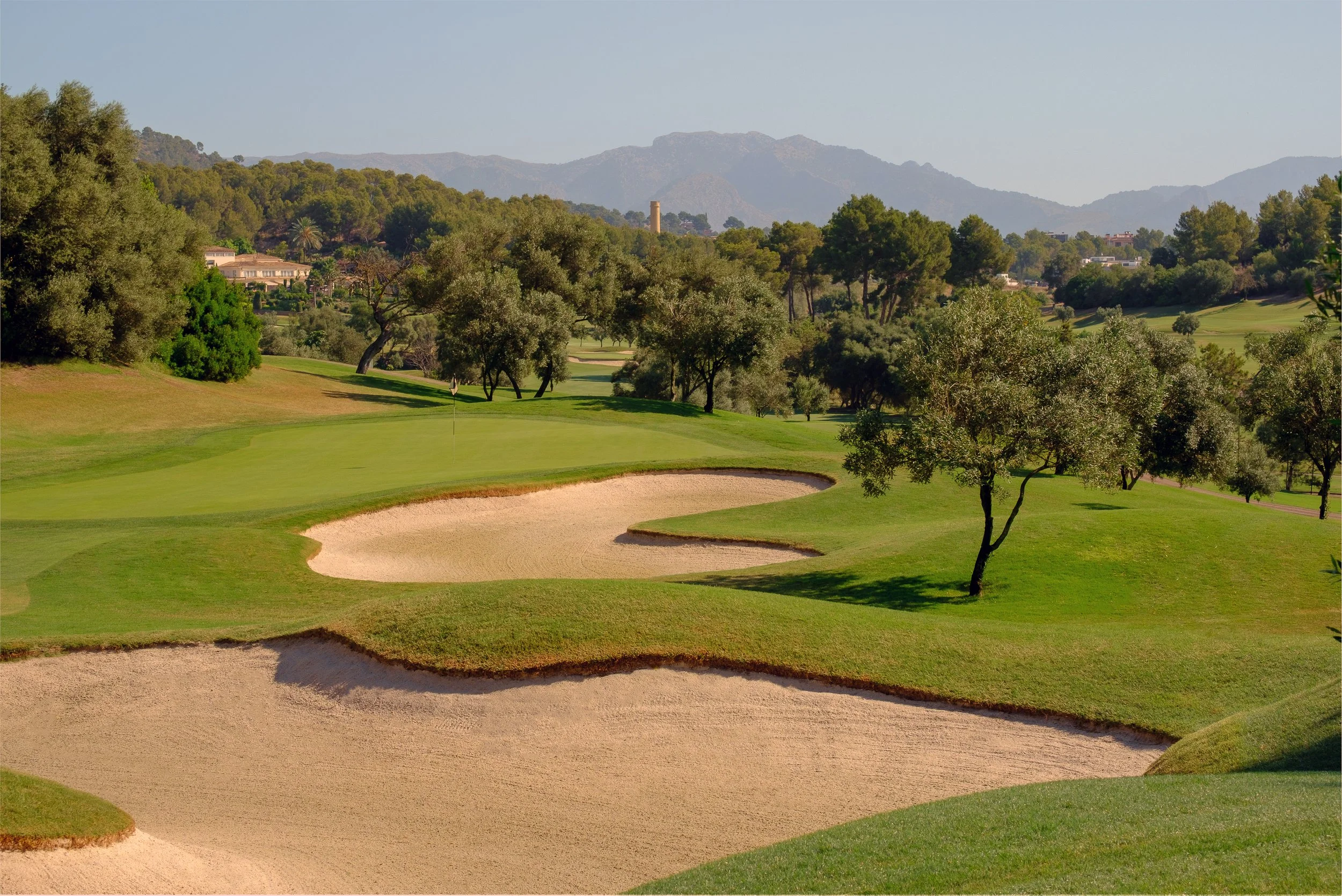 Campo de golf con búnkeres, árboles y colinas, en un día soleado con montañas en el horizonte.