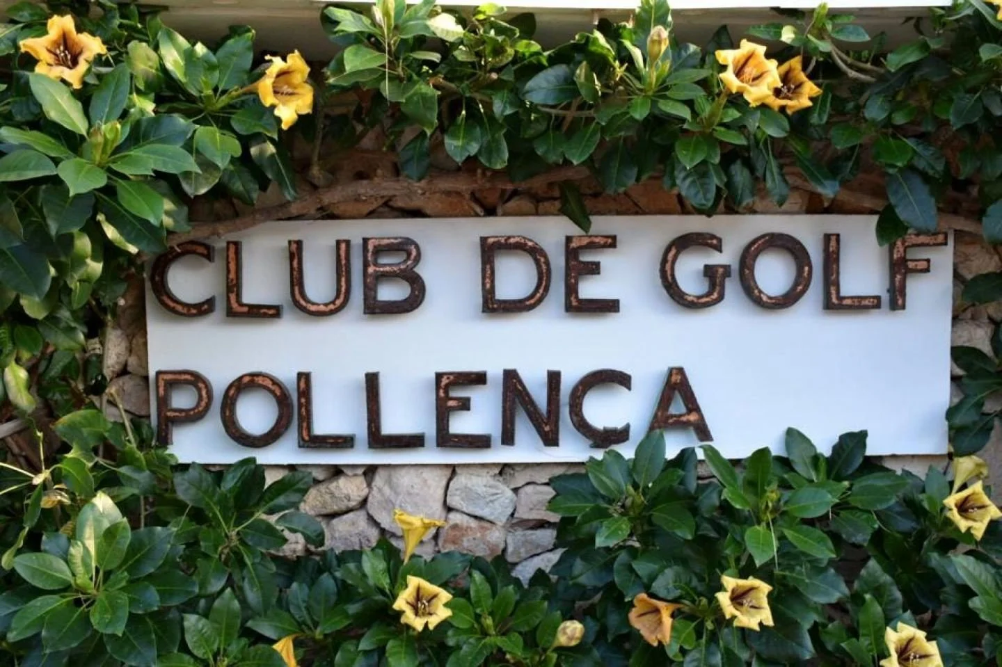 Placa en un muro de piedras con letras de metal que dice 'Club de Golf Pollença' rodeada de plantas verdes con flores amarillas.