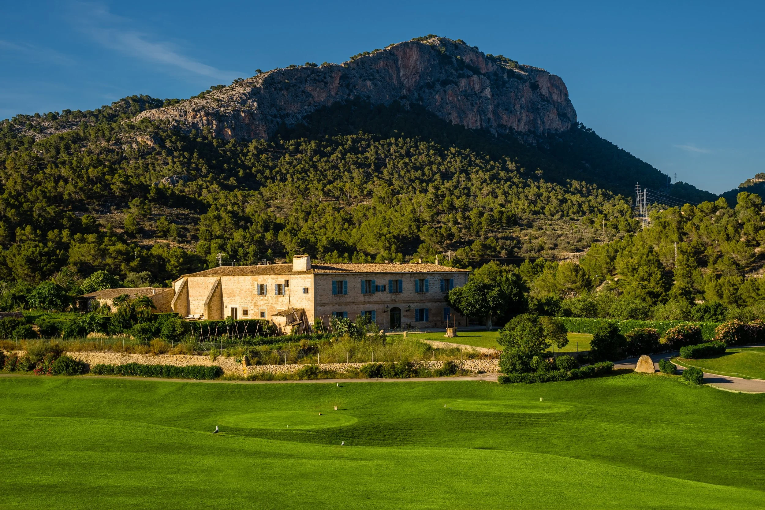 Vista de un campo de golf con césped bien cuidado, árboles y un edificio de piedra en el fondo, rodeado de montañas verdes bajo un cielo azul.