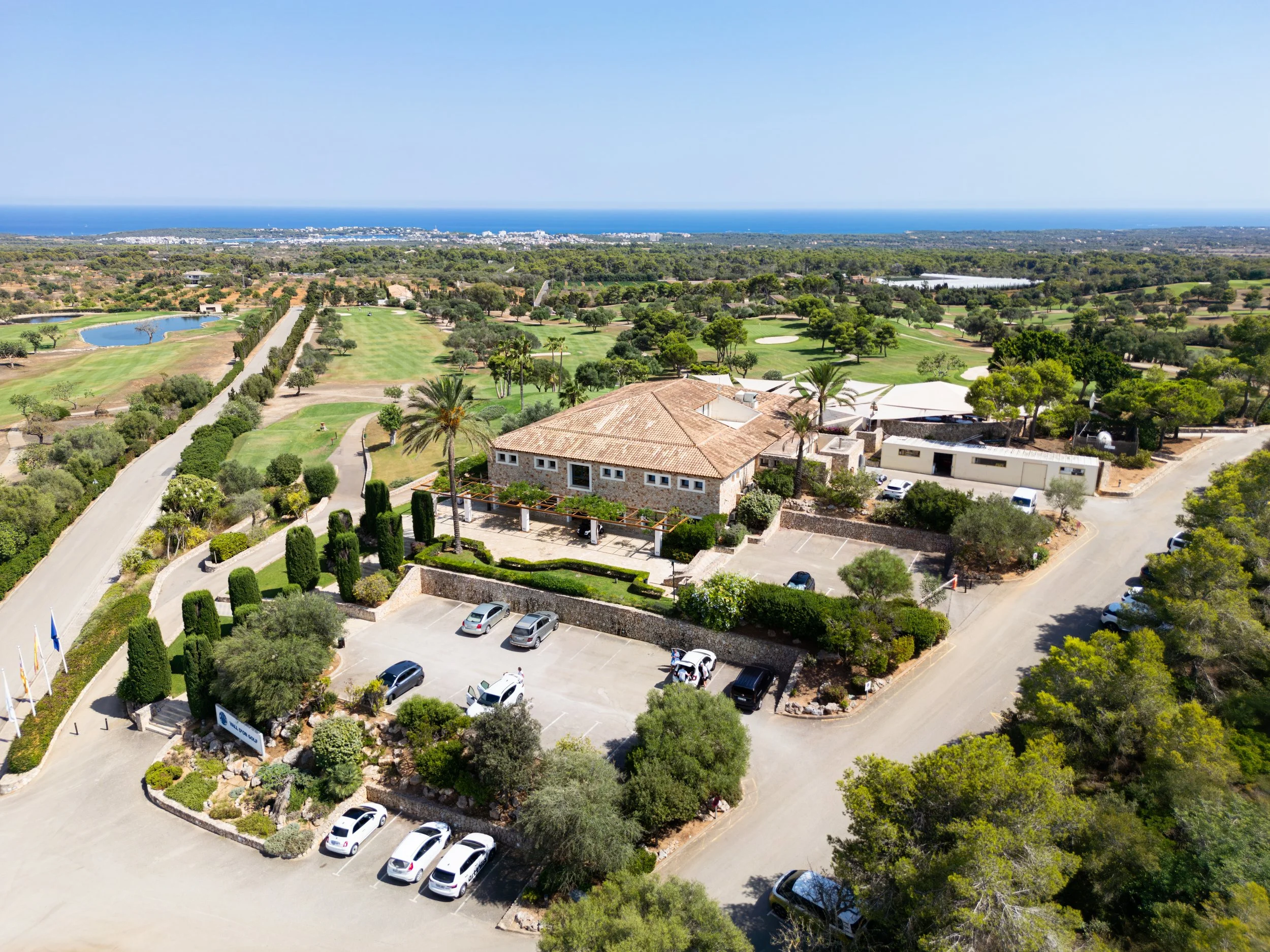 Vista aérea de un edificio rodeado de árboles y áreas verdes, con un campo de golf y el mar en el fondo.