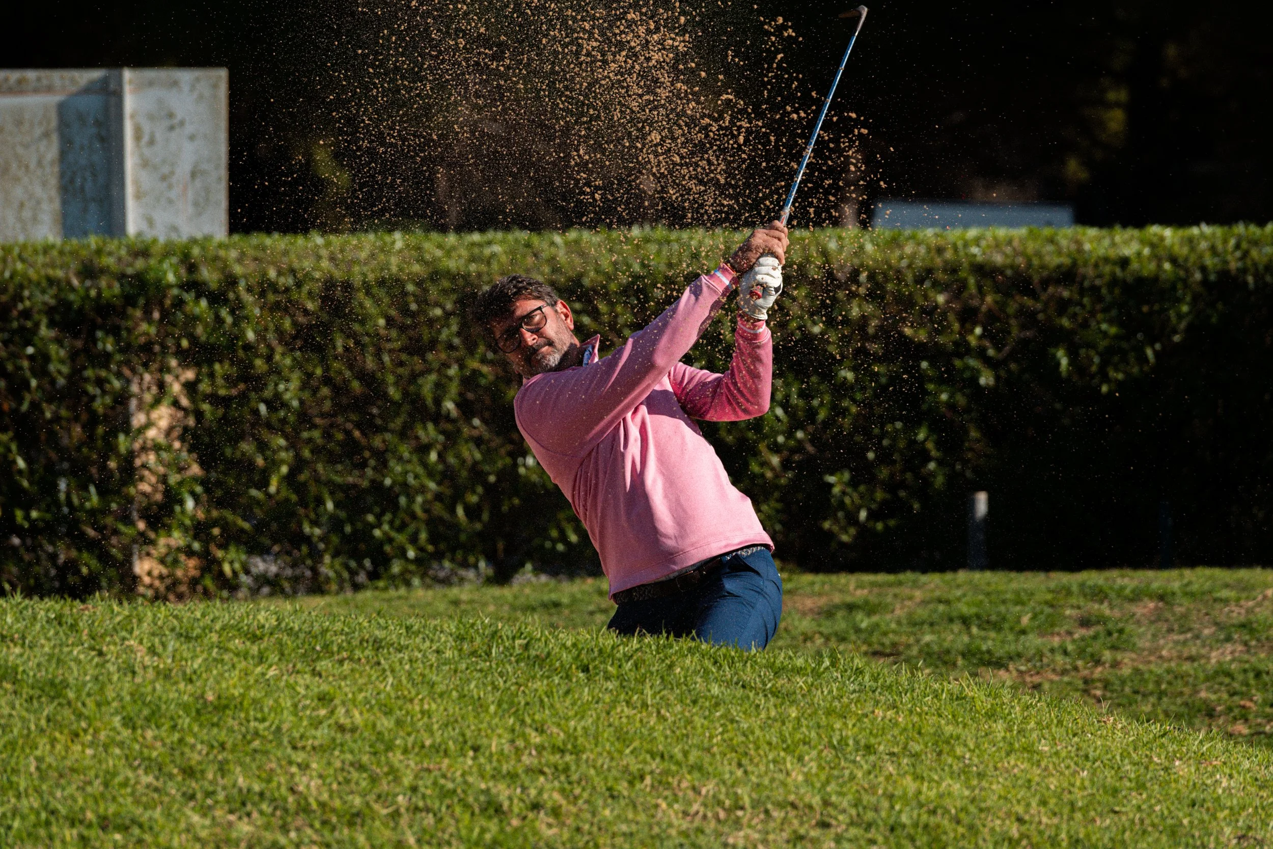 Hombre con barba y gafas, en un campo de césped, golpeando una pelota de golf con un palo, en medio de un swing.