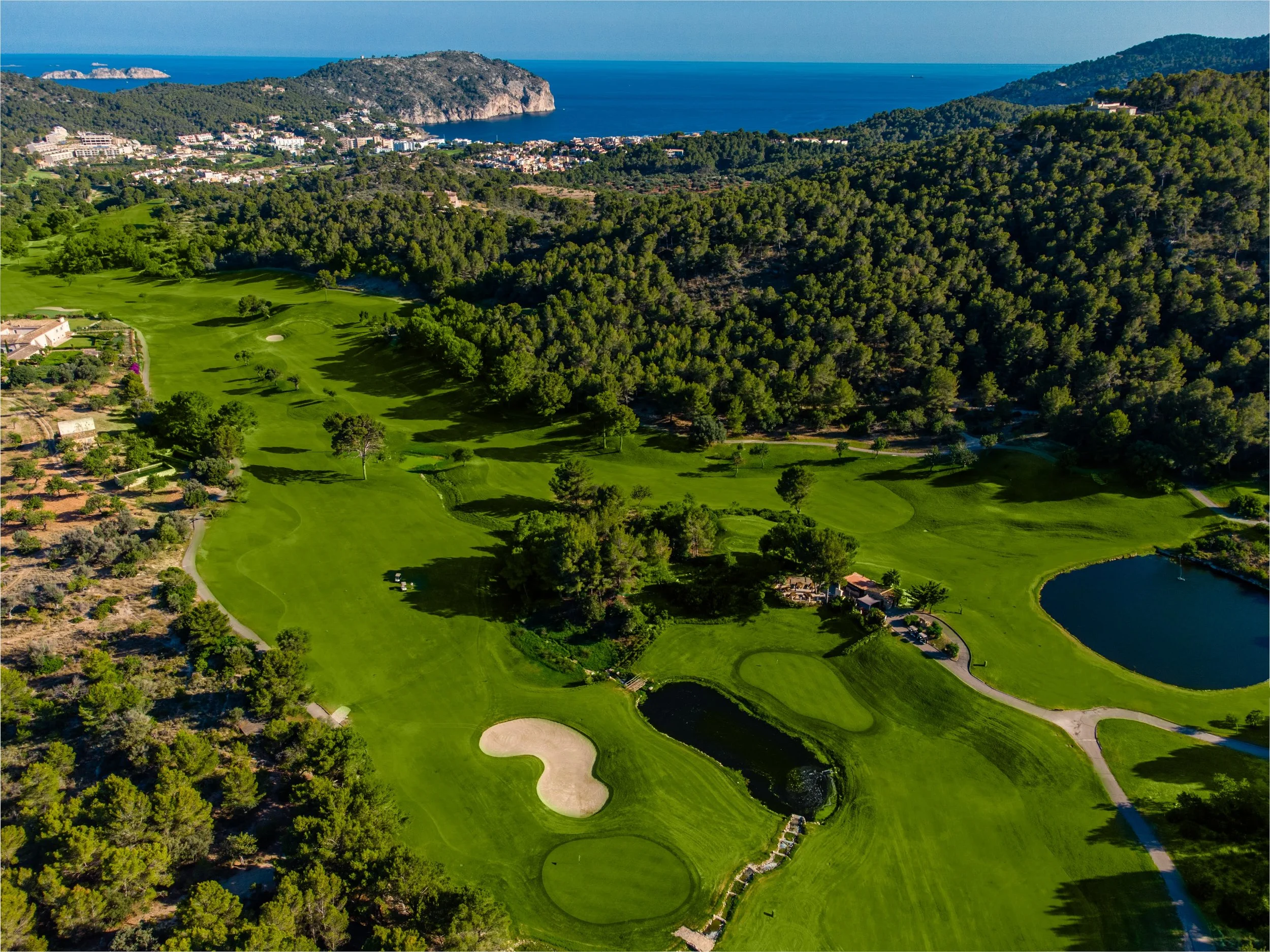 Vista de un campo de golf rodeado de árboles y con un lago y el mar en el fondo.