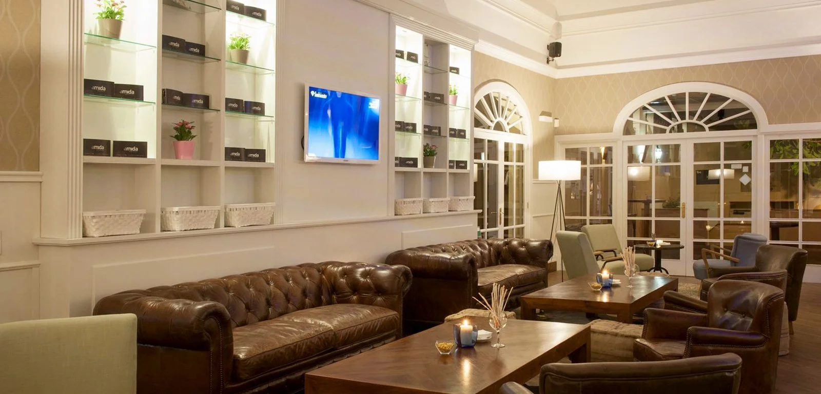 Sala de estar con sillones de cuero, sillón de tela, mesas de madera, lámpara de pie, televisión en la pared, ventanas grandes con vista a la calle, decorada con plantas y velas.