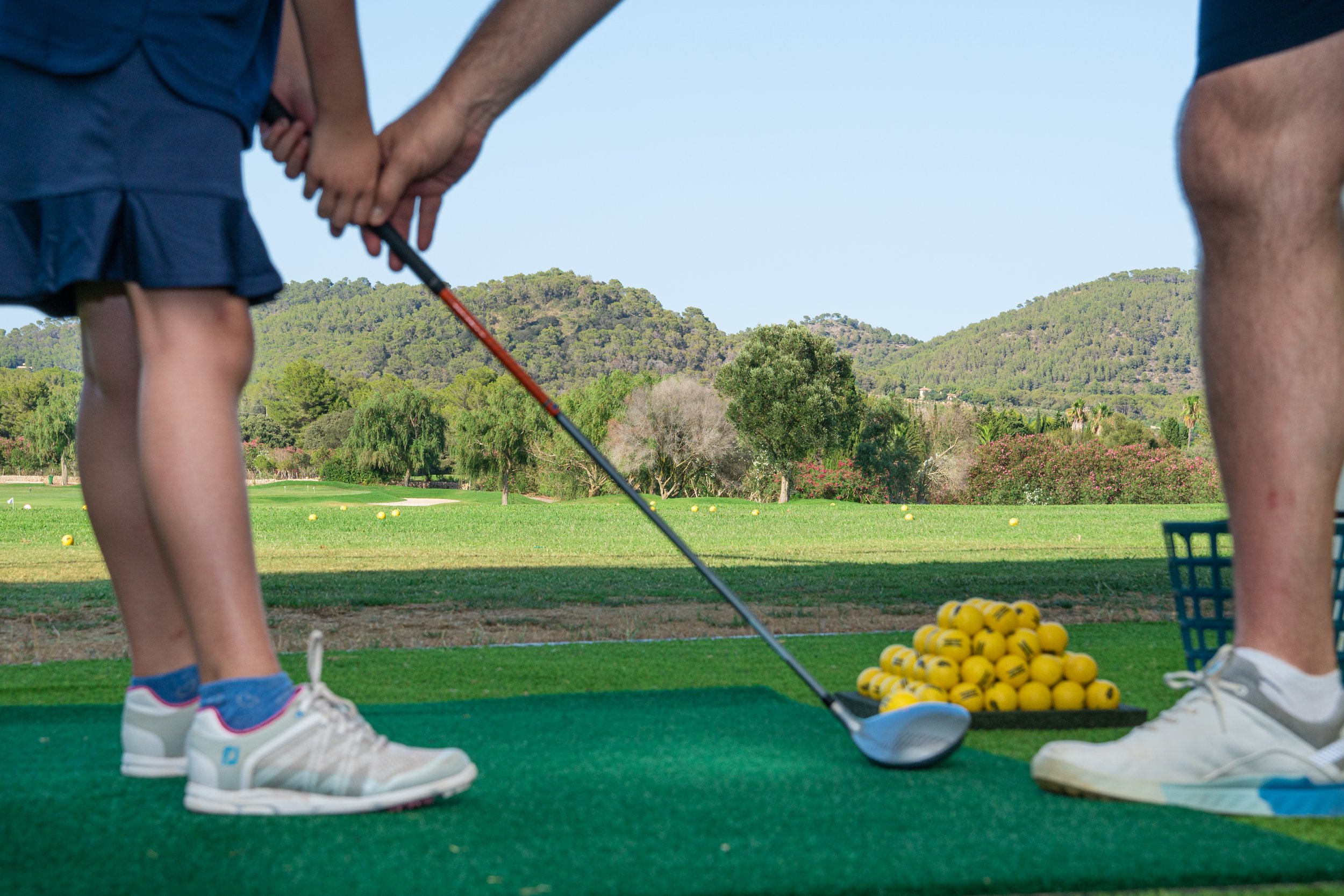 Un niño y un adulto en un campo de golf preparándose para jugar, con montones de pelotas amarillas y un paisaje de montañas y árboles en el fondo.
