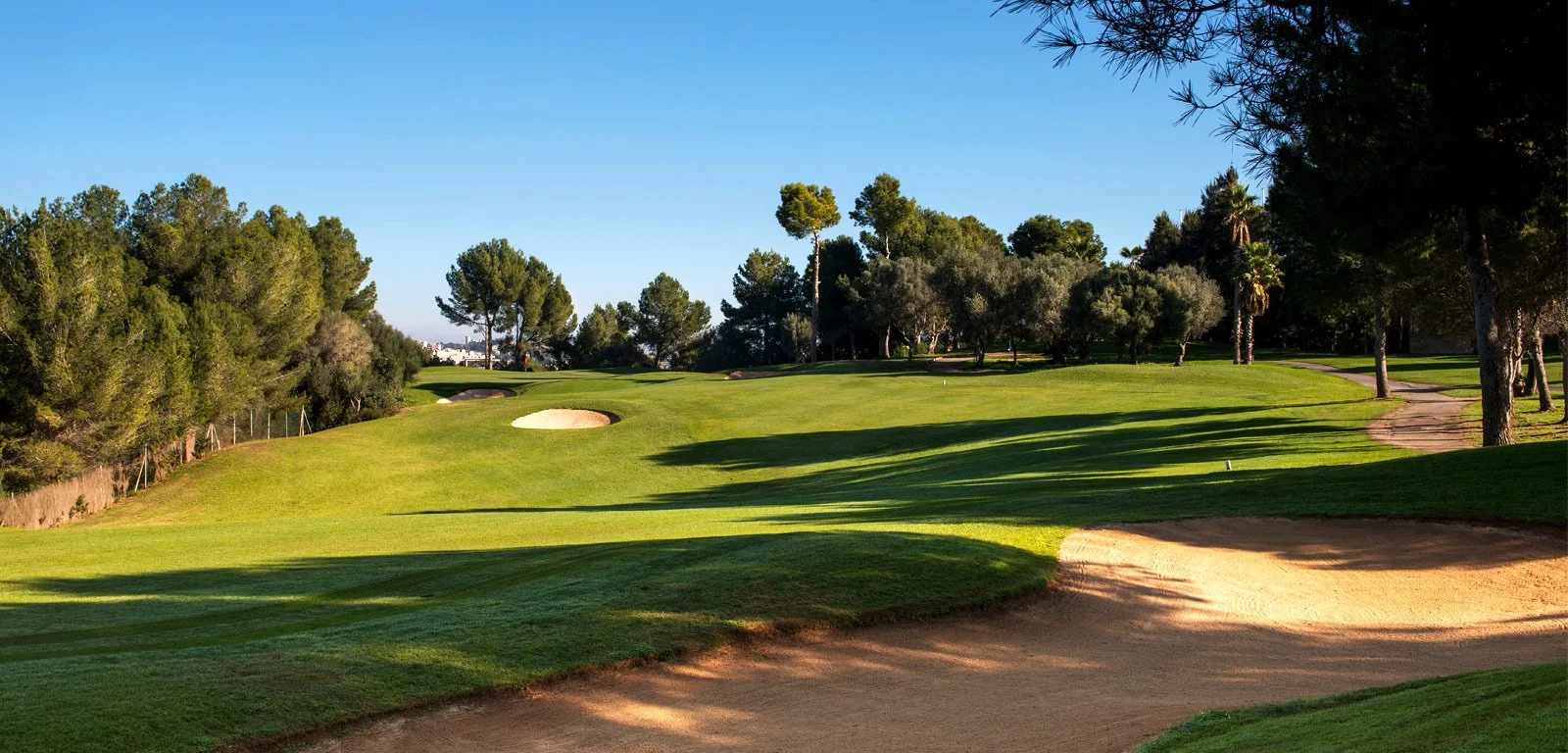 Campo de golf con césped verde, algunos obstáculos de arena, árboles y camino de tierra en un día soleado y despejado.