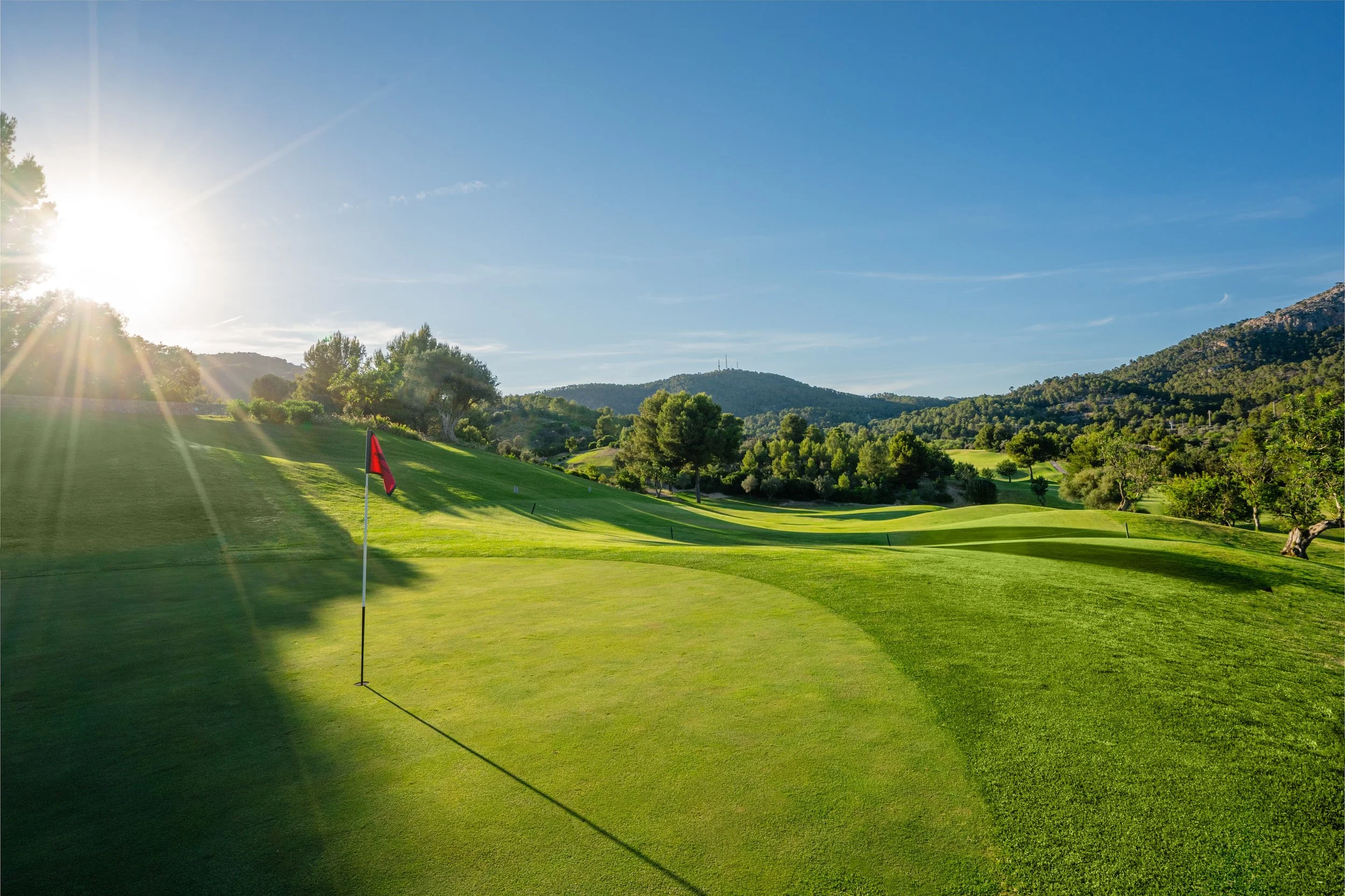 Campo de golf con césped verde, bandera roja, árboles y colinas en el fondo, sol brillante y cielo azul