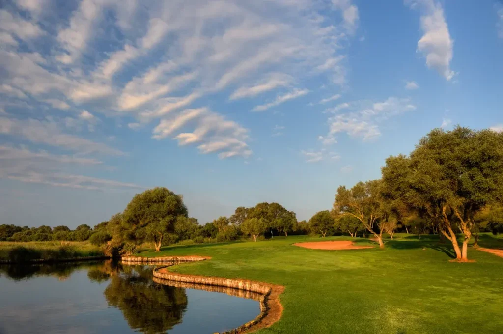 Un campo de golf con árboles y un estanque, cielo despejado con algunas nubes.