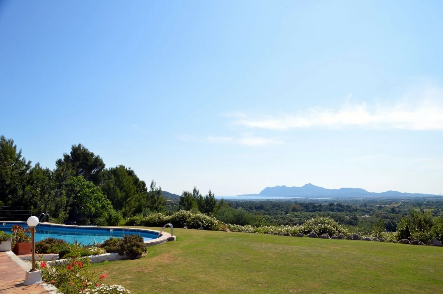 Vista de un jardín con piscina en primer plano, rodeada de plantas y flores, con un campo y montañas al fondo bajo un cielo despejado