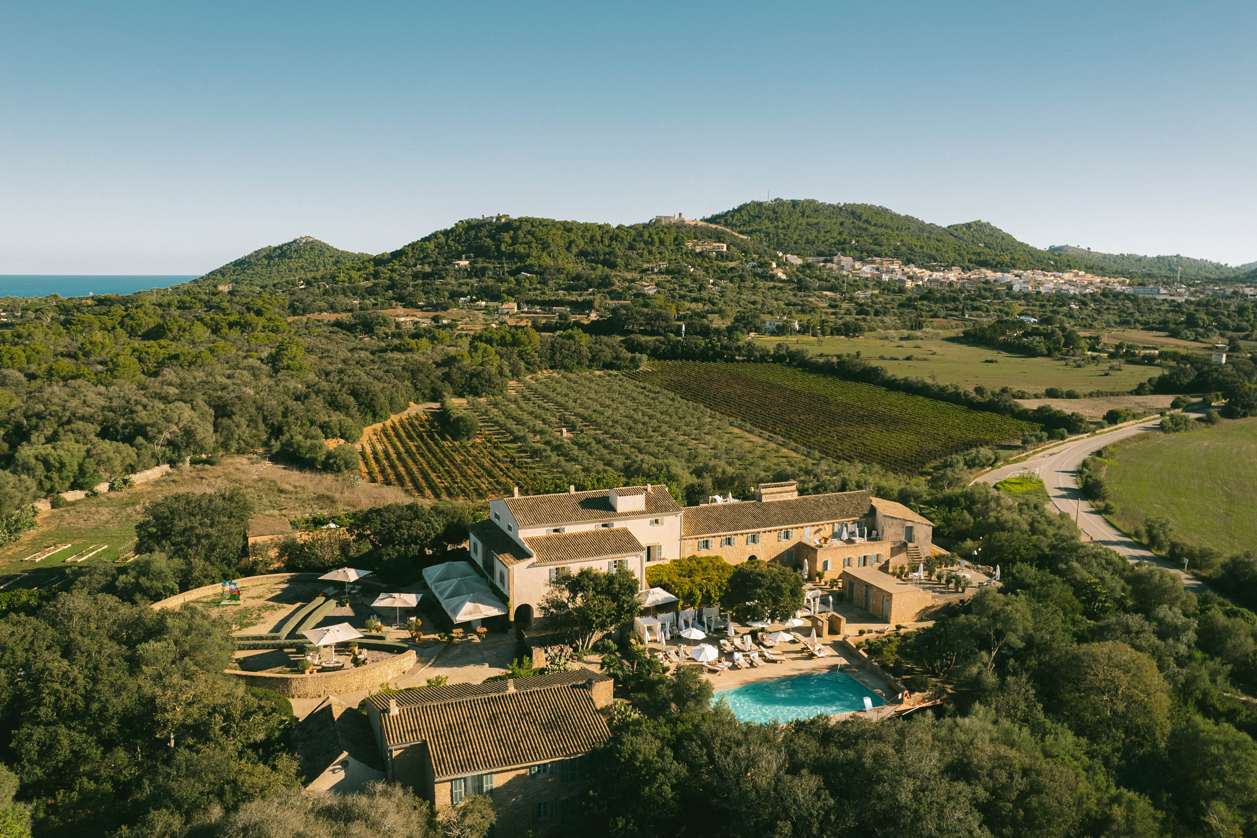 Paisaje con colinas verdes, casas y un área de piscina con sombrillas en primer plano, y el mar visible en la distancia bajo un cielo despejado.