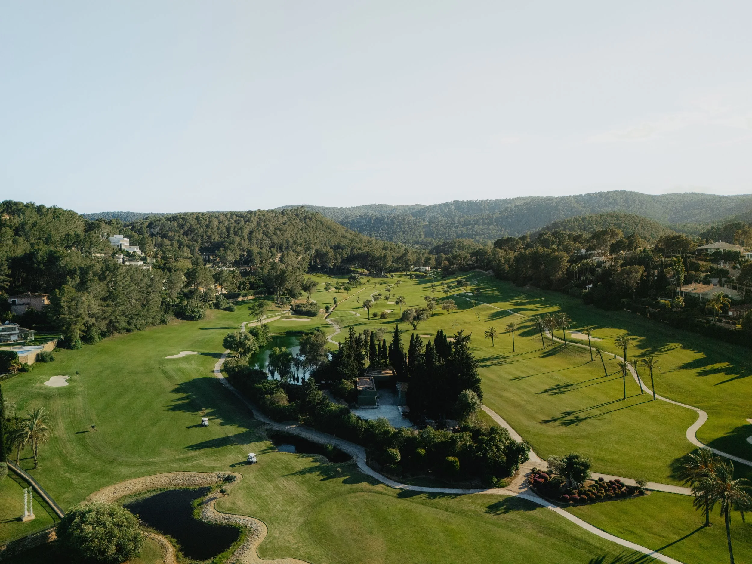 Vista aérea de un campo de golf rodeado de árboles y colinas, con senderos pavimentados y una pequeña laguna en el centro.