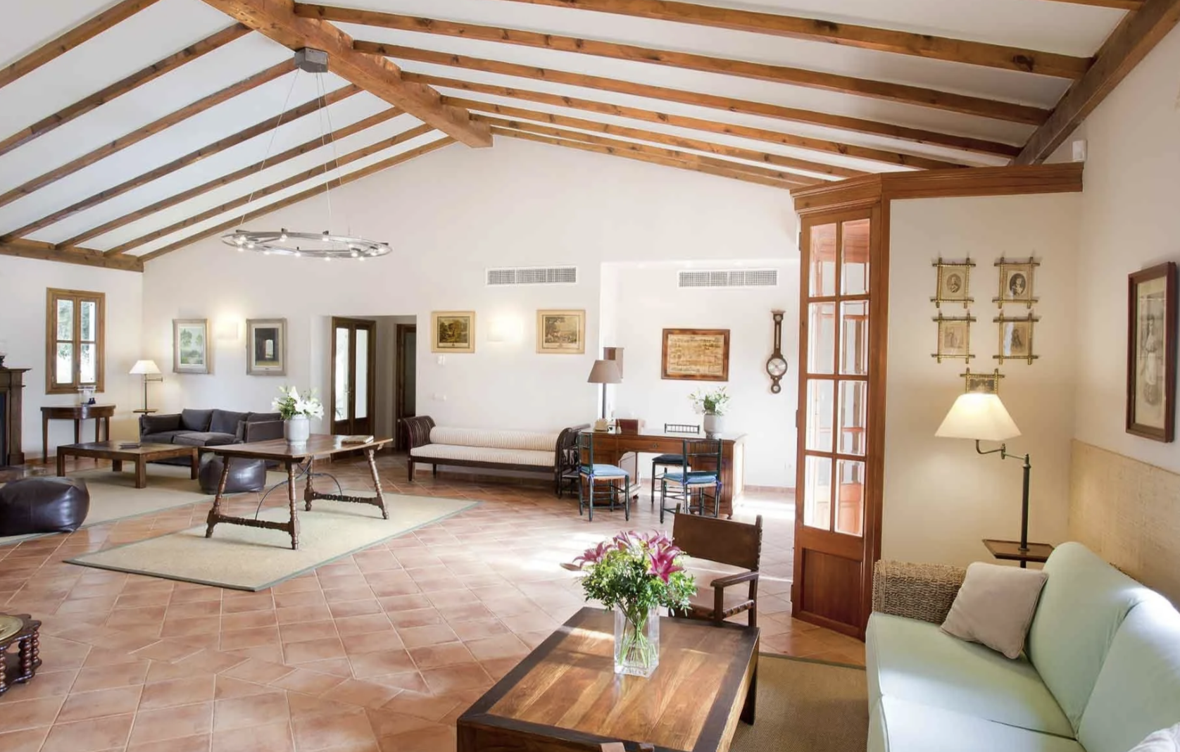 Sala de estar con techo a dos aguas y vigas de madera visible, muebles de madera y tapiz, varias lámparas, ventanas y cuadros en las paredes, plantas decorativas, suelo de baldosas y espacio amplio y luminoso.