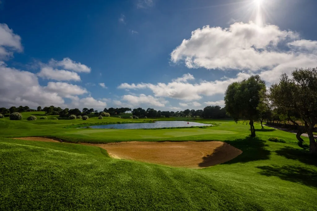 Campo de golf con arboles, un estanque y un bunker en un día soleado con nubes en el cielo.