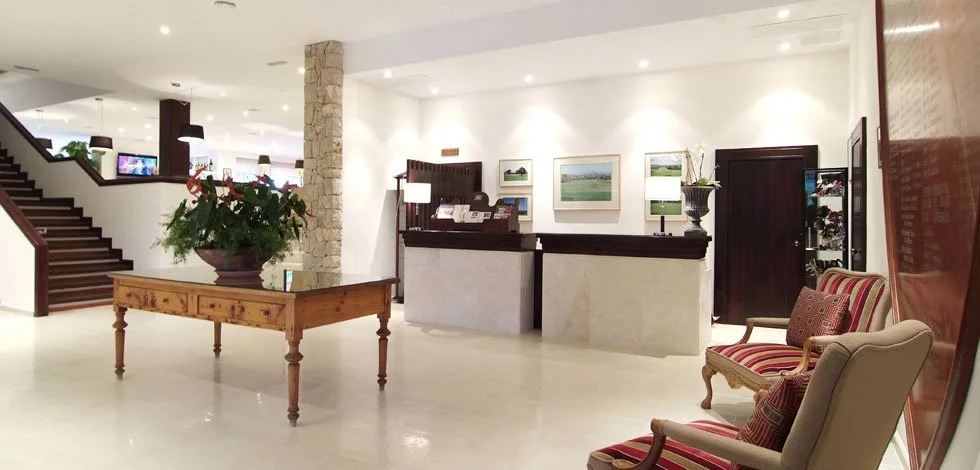 Vestíbulo de un hotel con una mesa de madera con una planta en el centro, paredes blancas decoradas con cuadros, una recepción con escritorio, y varias sillas tapizadas con franjas rojas y beige.