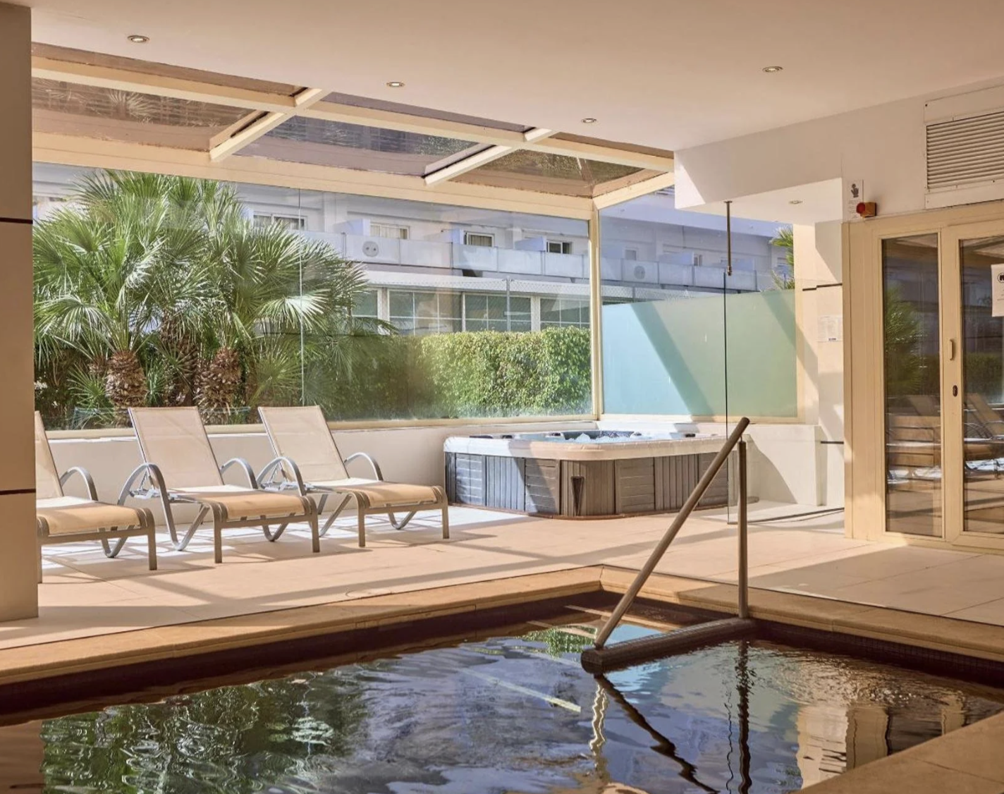 Área de descanso en un spa con sillones reclinables, jacuzzi y vista a plantas y edificio en el fondo.