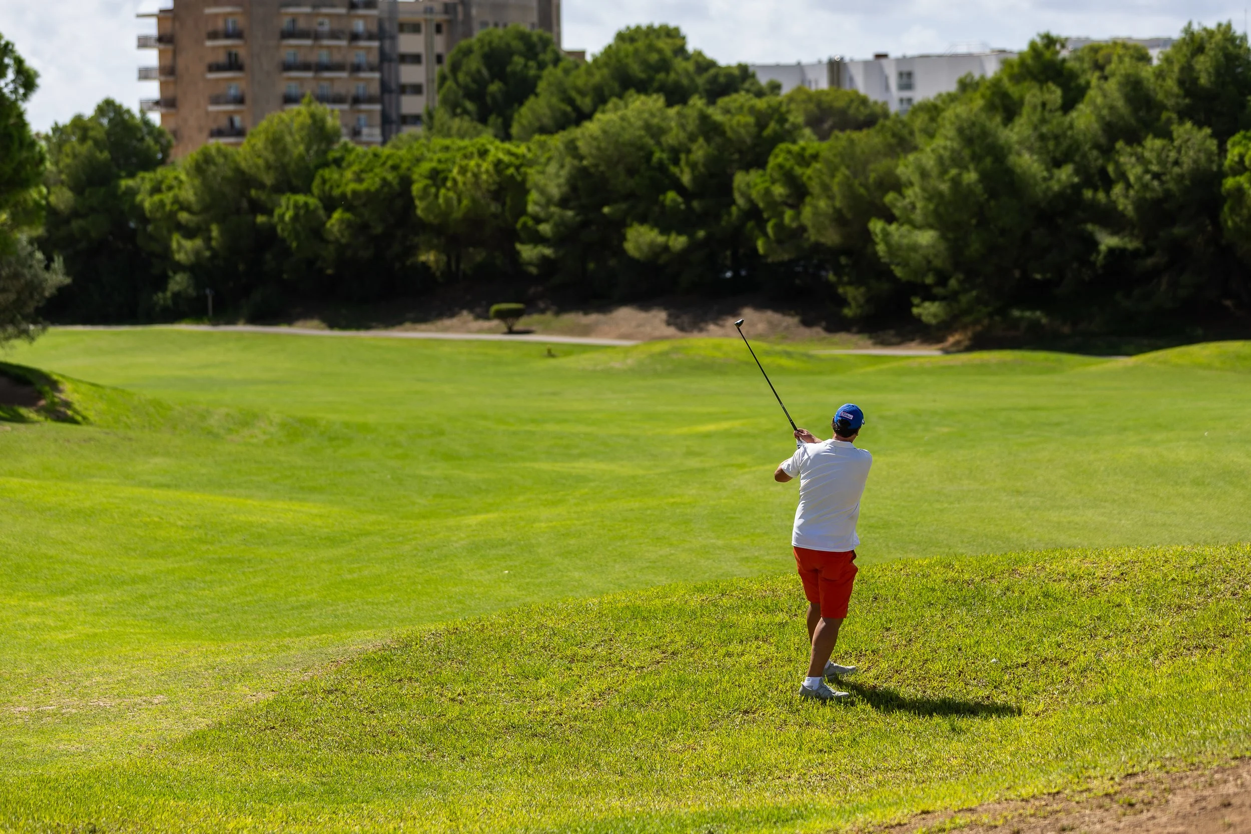 Persona jugando golf en un campo con césped verde y árboles, con edificios en el fondo.