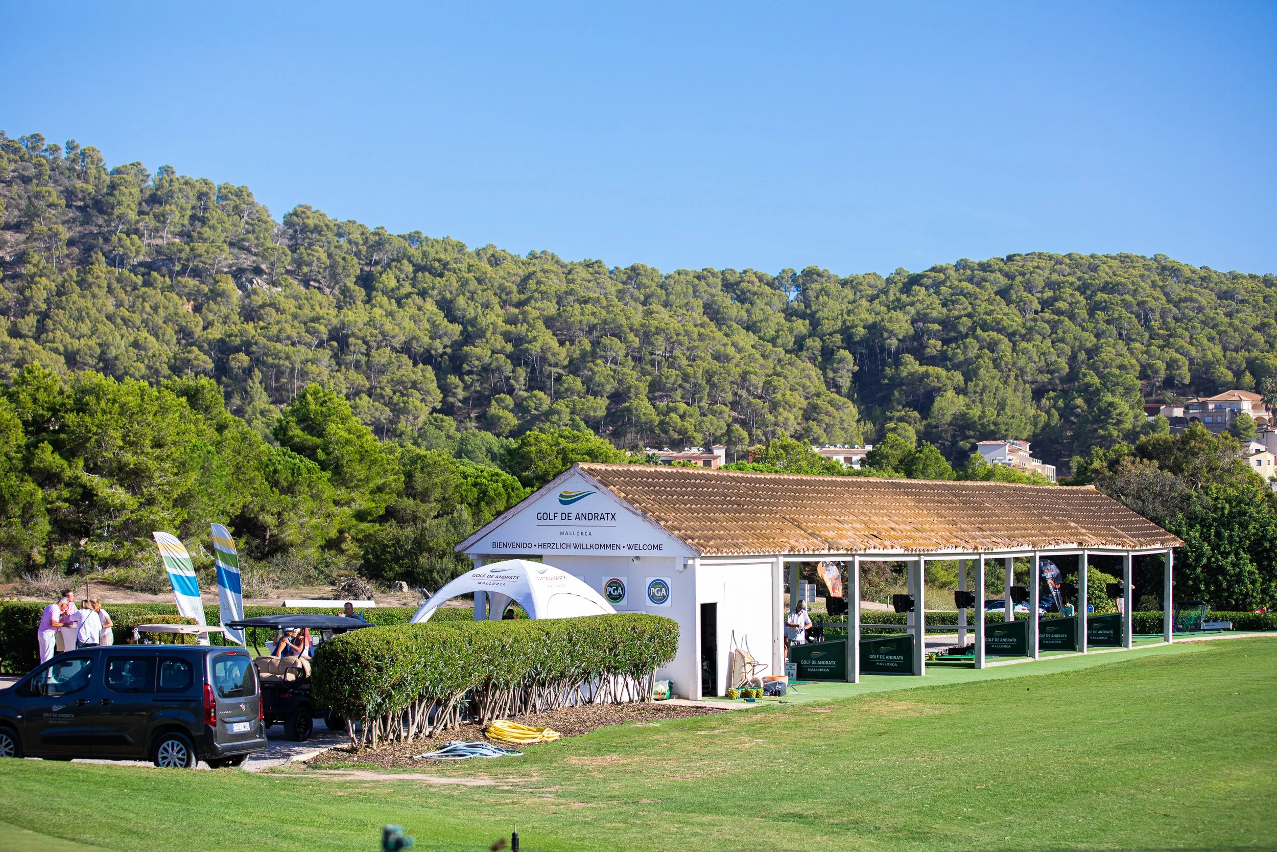 Vista de una academia de golf al aire libre con un edificio blanco y toldos, rodeada de césped y árboles verdes, con colinas y casas en el fondo, y el cielo despejado.