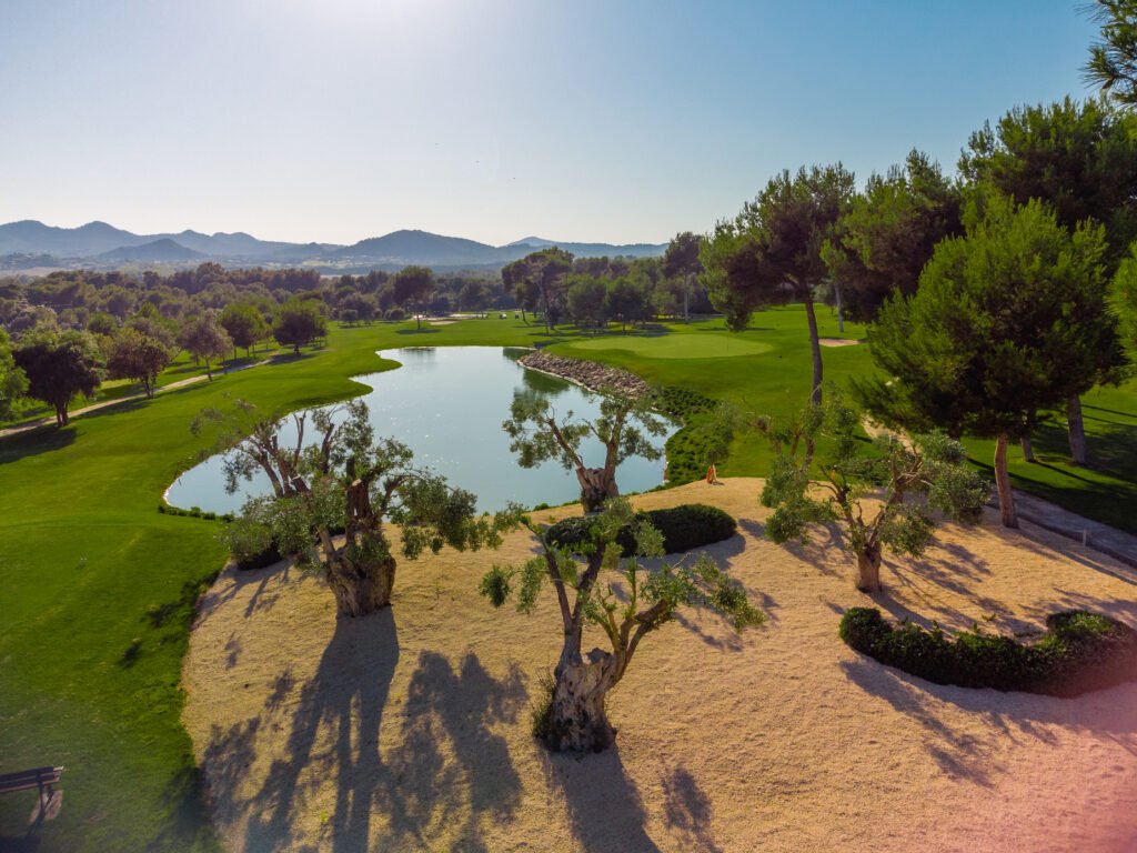Campos de golf con árboles, estanque y montañas de fondo en un día soleado.