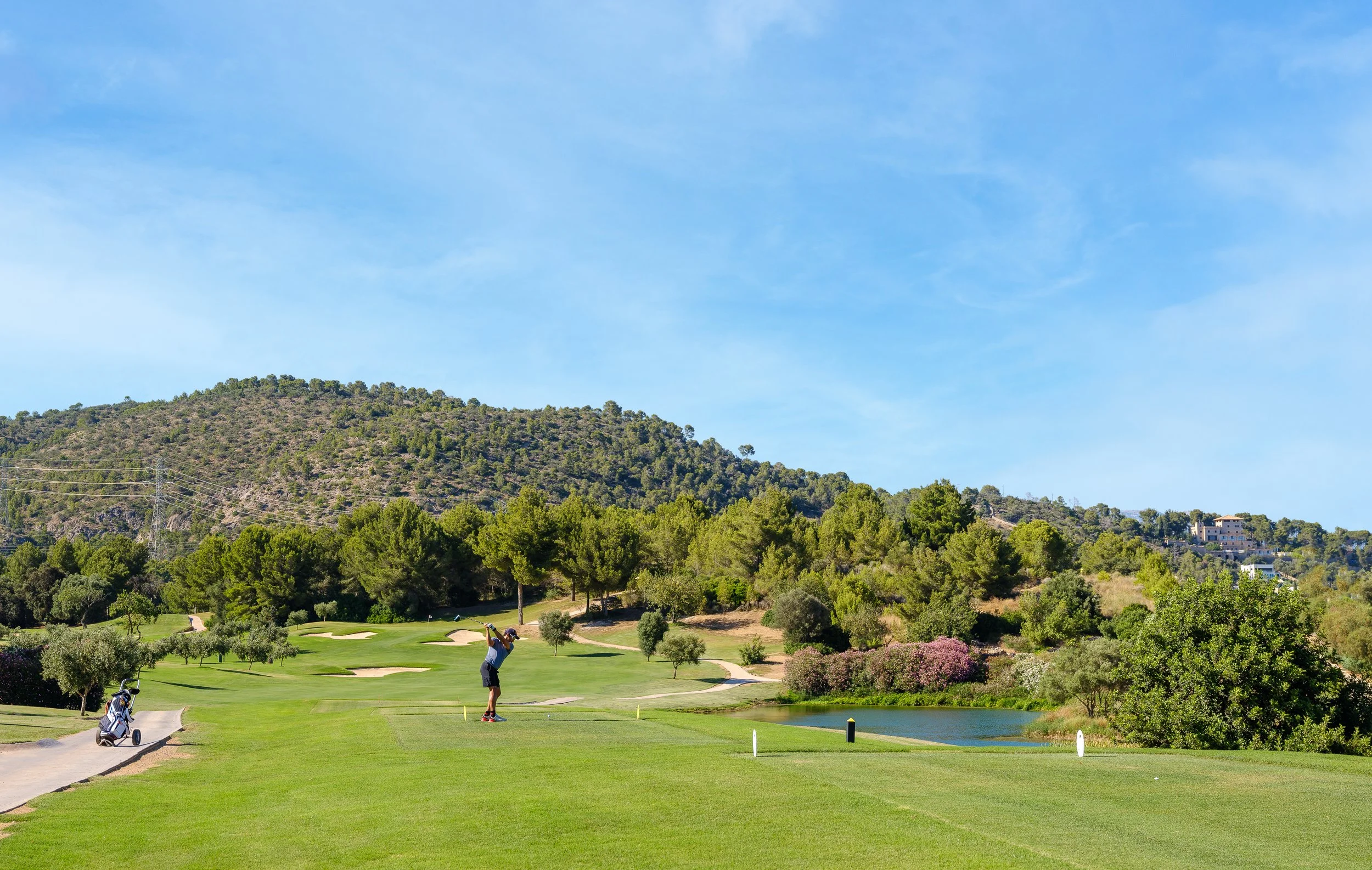 Un campo de golf con un jugador preparado para hacer un golpe, un carrito de golf y un lago, rodeado de árboles y colinas bajo un cielo azul.