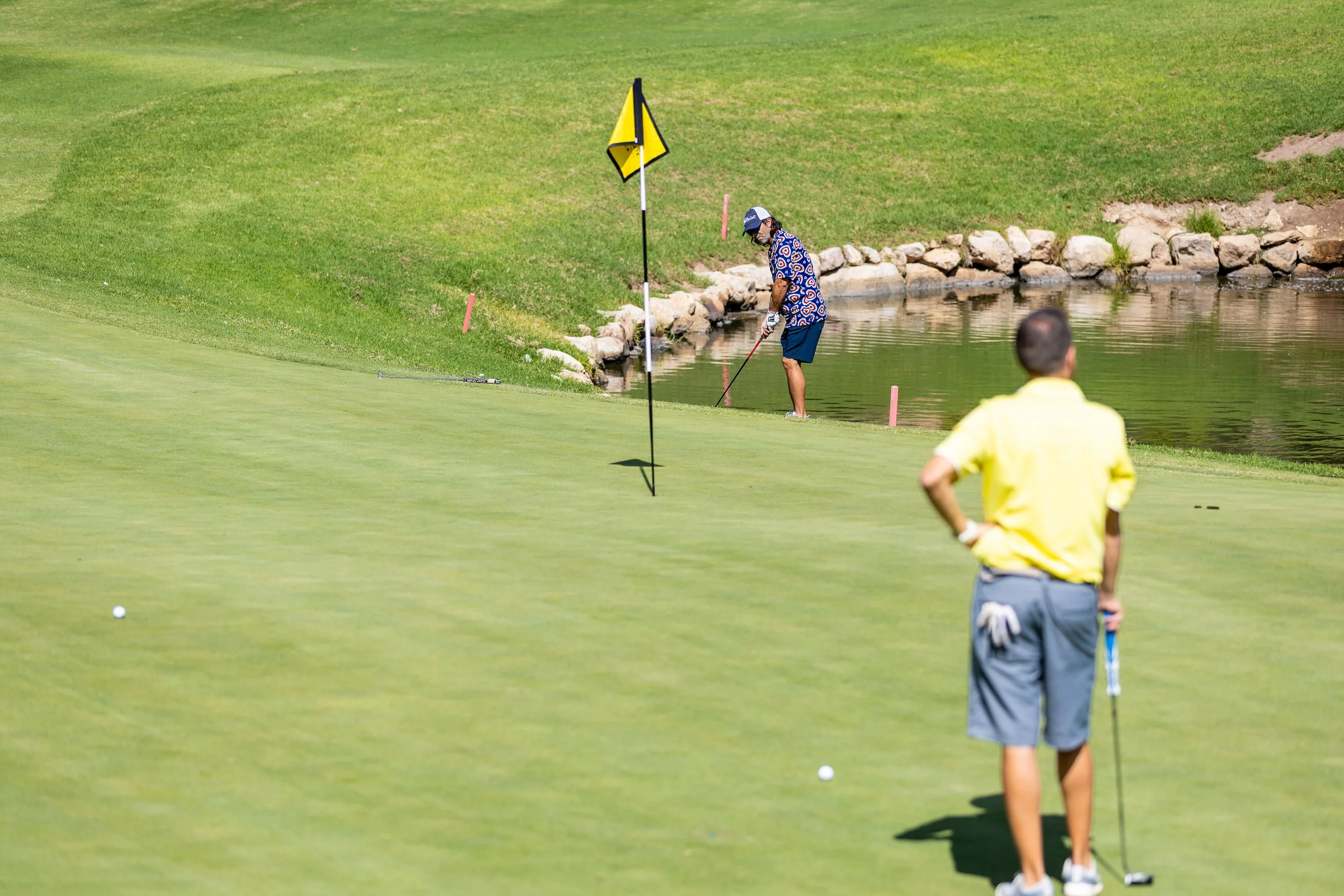 Jugador de golf en un campo cercano a un lago, preparado para hacer un tiro, con otro jugador observando desde cerca.
