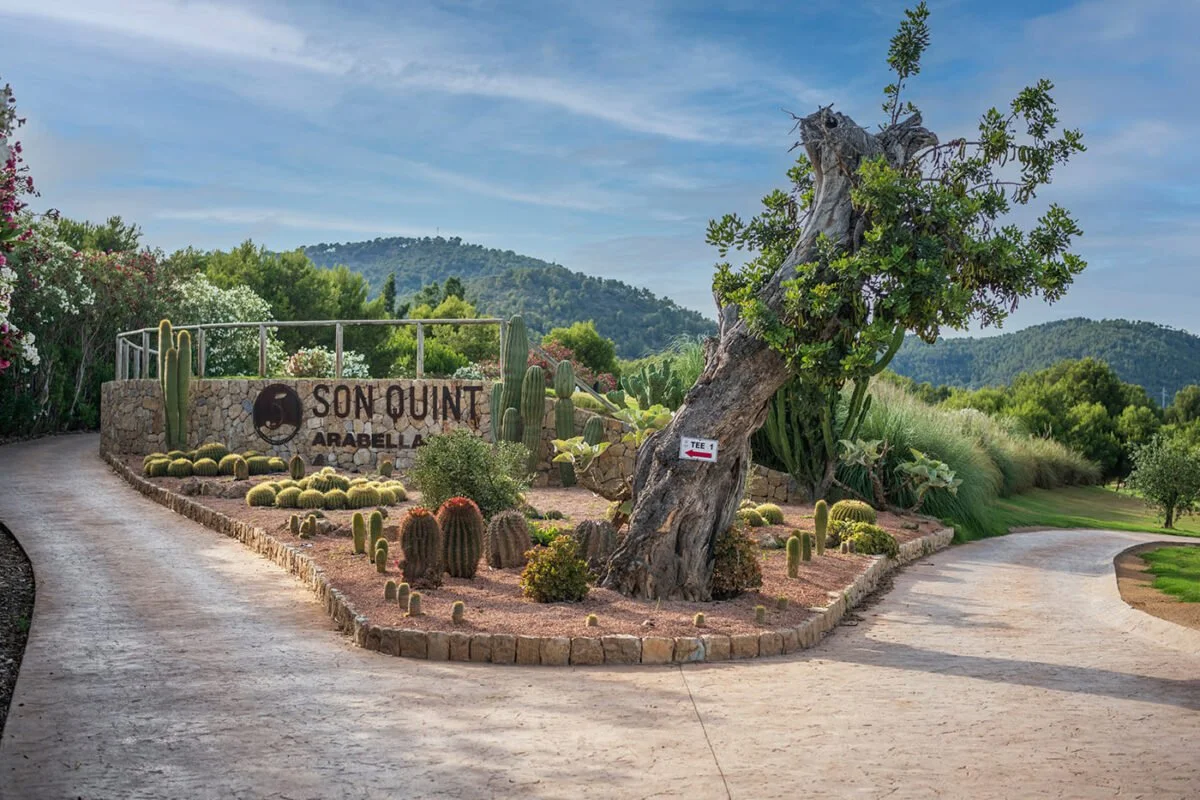 Entrada a un campo de golf llamado 'Son Quint Arabelle', rodeada de cactus y plantas en un paisaje con colinas y cielo despejado.
