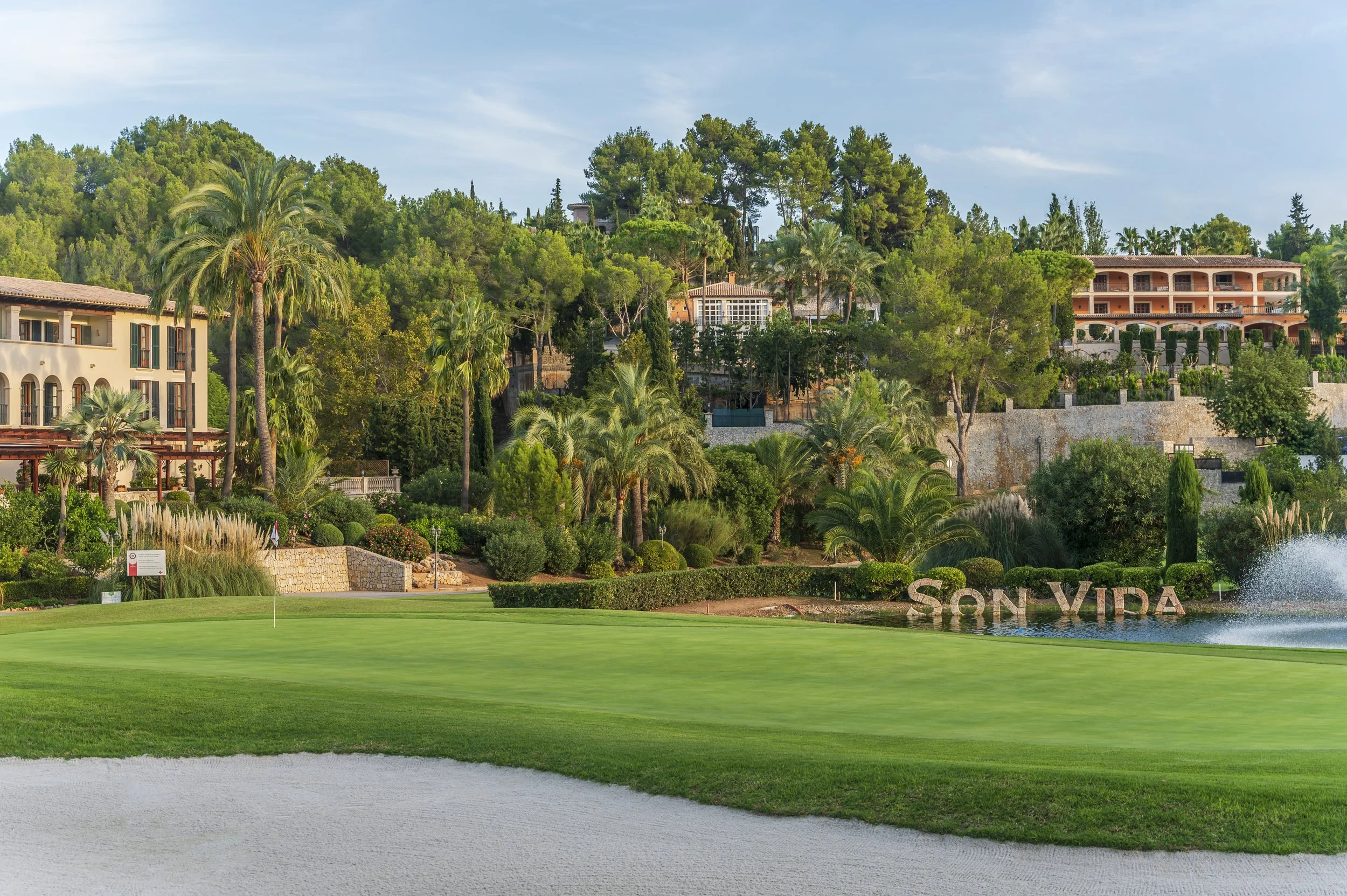 Campo de golf con césped bien cuidado, arena en el bunker, árboles de palmeras y otros tipos de árboles, casas y terrazas en la colina rodeada de vegetación, señal que dice "Son Vida" y fuente de agua en el fondo.