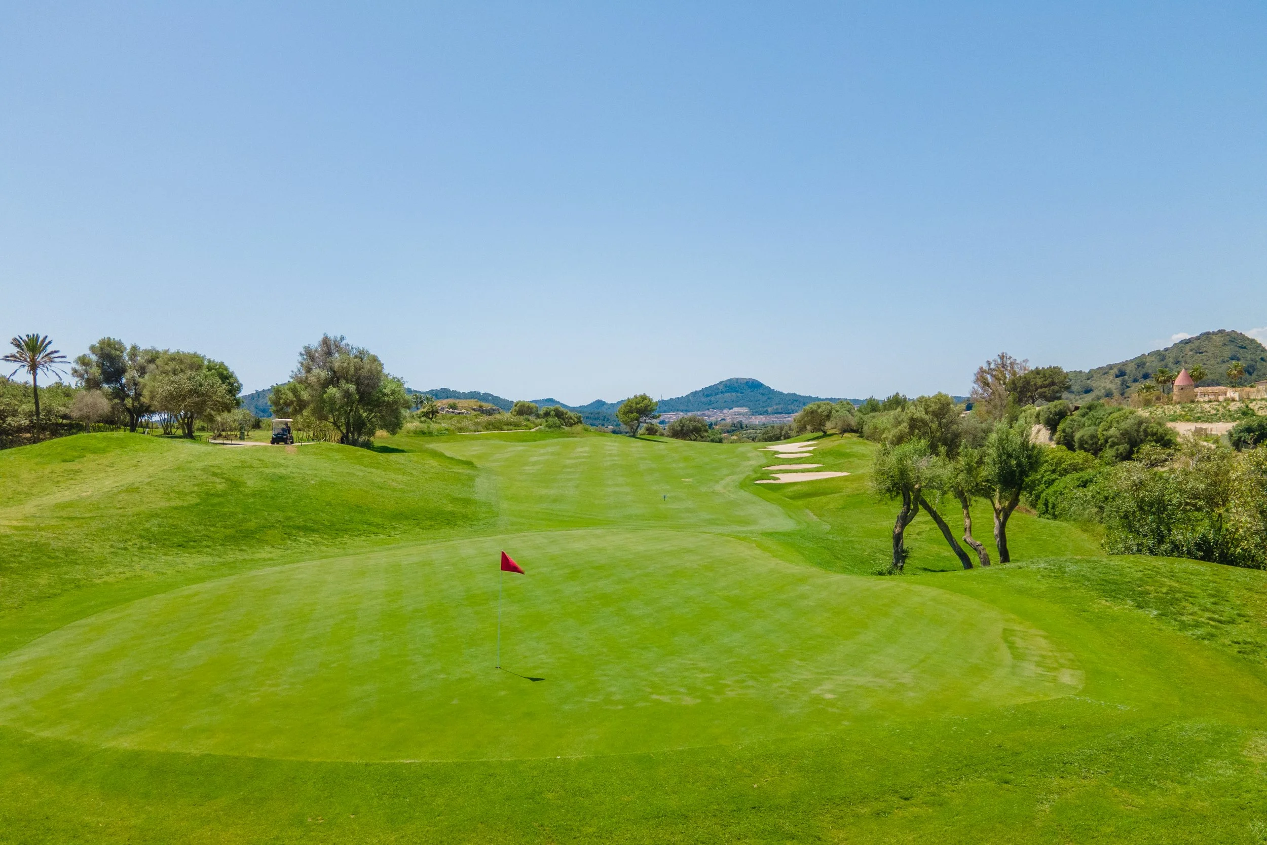 Campo de golf con césped verde, árboles y montañas en el fondo, bajo un cielo despejado.
