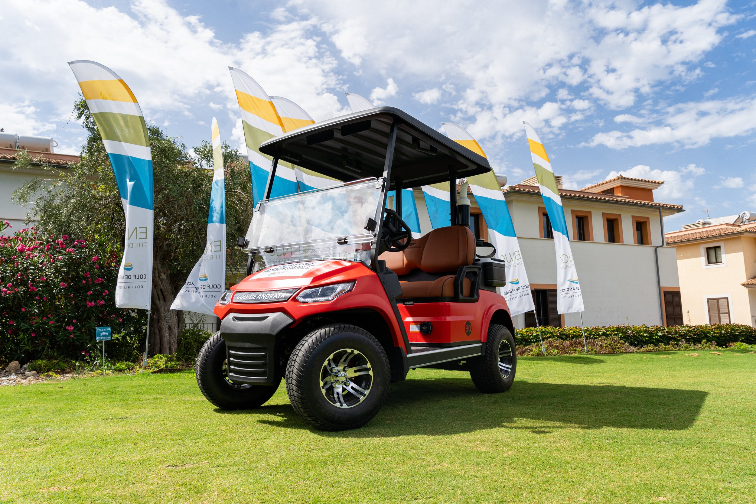 Carrito de golf de color rojo en un campo de césped con banderas blancas, azules y verdes y casas en el fondo bajo un cielo con nubes.