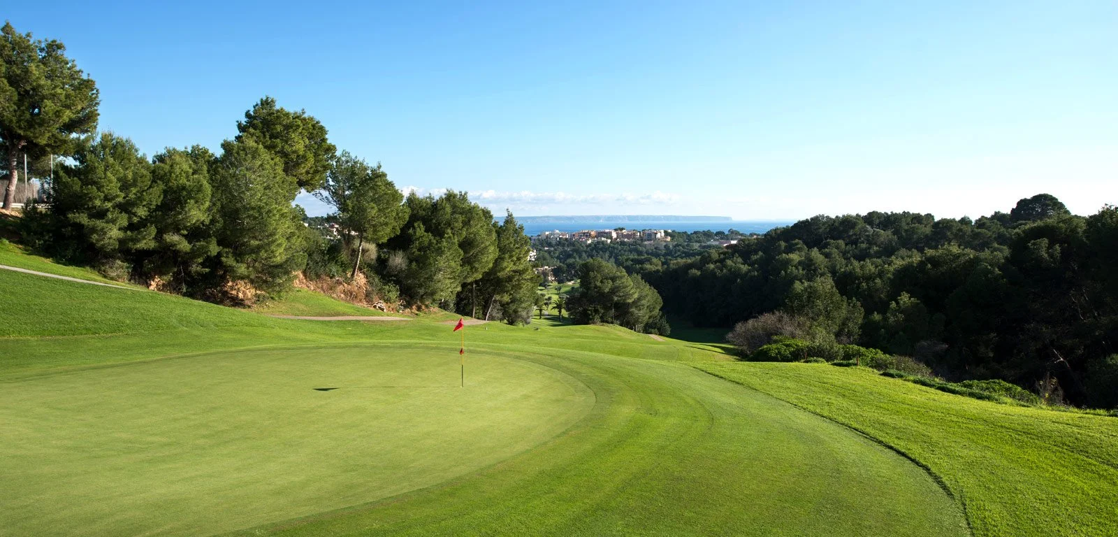 Un campo de golf con un hoyo y bandera, rodeado de árboles verdes y colinas, con un cielo despejado y vista al fondo del mar y la ciudad.